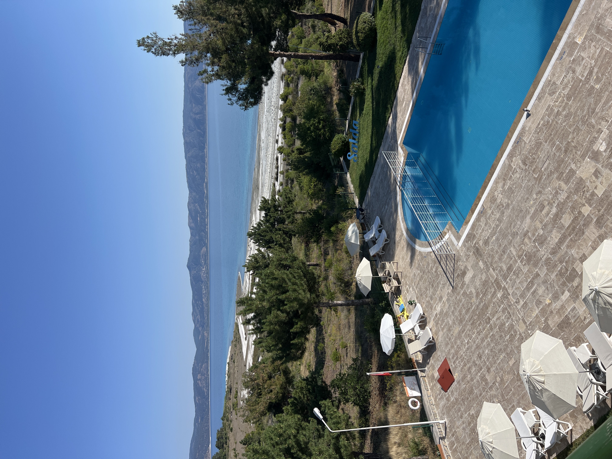 This image depicts a poolside scene at a resort in Yeşilova, Turkey, overlooking Lake Salda. The foreground features a swimming pool with bright blue water and a white railing. Surrounding the pool is a tiled patio with several lounge chairs and white umbrellas. Beyond the patio, a grassy area has a blue sign that reads "Salda". A cluster of pine trees separates the resort area from the natural landscape. The background is dominated by the expansive, turquoise waters of Lake Salda, with a white, sandy shore and distant, rolling hills under a clear blue sky. The scene is tranquil and suggests a pleasant day at a lakeside resort.