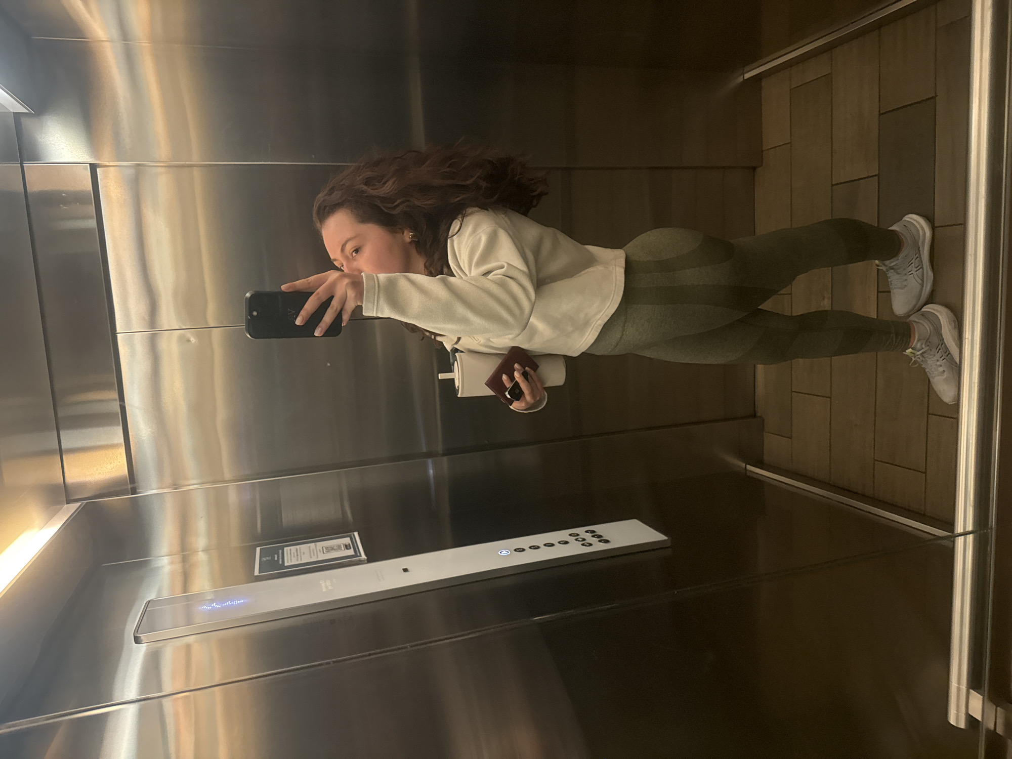 A young woman with curly brown hair and wearing olive-green leggings, a white sweatshirt, and grey sneakers is taking a photo of herself in a mirrored elevator. She is holding a black smartphone in her left hand. In her right hand, she is holding a white cup and a small brown object. The elevator has a stainless steel interior with a row of buttons along the wall. The floor is tiled in brown. The location of the elevator may be in the city of Mexico City, Mexico.