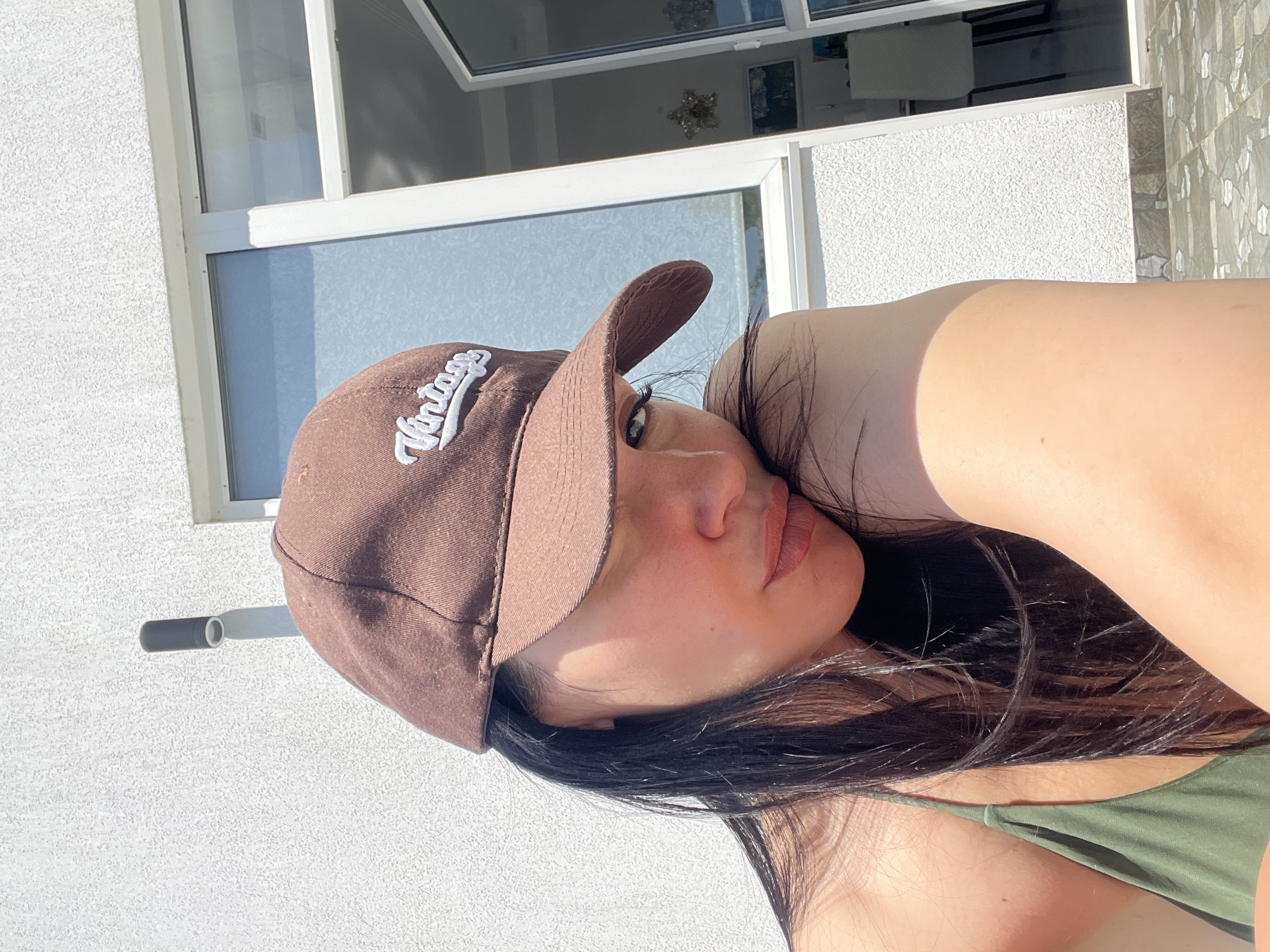 The image captures a woman in a close-up, slightly tilted shot, suggesting a relaxed pose outdoors in Baranovka, Russia. She wears a brown baseball cap with the word "Vintage" embroidered in white script on the front. Her long, dark hair frames her face, and she looks towards the camera with a calm expression, her lips slightly parted. She is wearing a green top, partially visible in the lower right corner. One of her hands is gently placed near her neck or shoulder.
The background features a textured white wall, possibly stucco, illuminated by bright sunlight which casts distinct shadows. Above and to the left, white-framed windows reflect the bright sky or an interior space, with some reflections showing what appears to be a decorative star or light fixture. A section of light-colored, patterned tiles is visible on the far right of the background. The strong, direct sunlight indicates it is daytime.