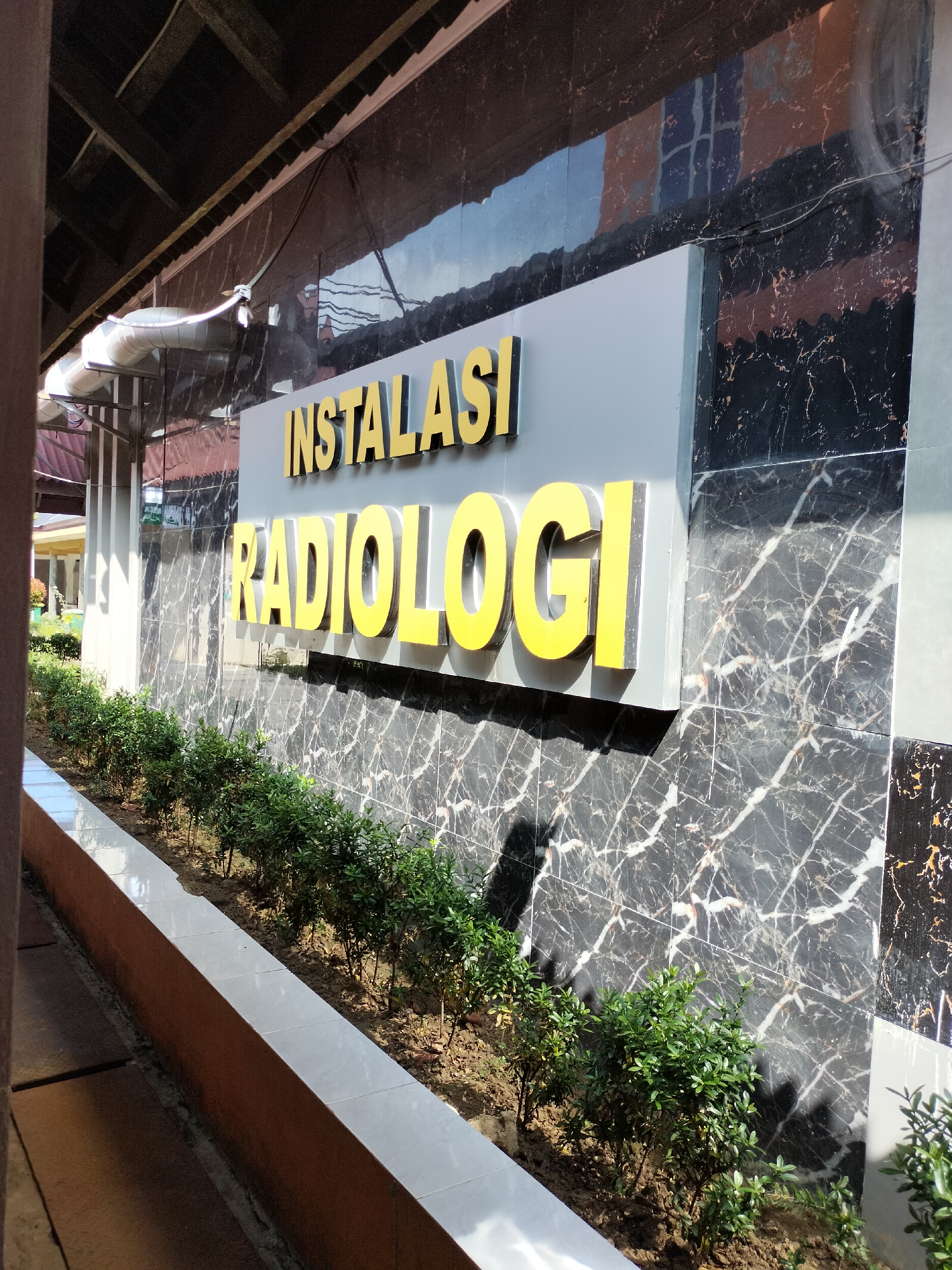 The image shows the facade of a building with a sign that reads "INSTALASI RADIOLOGI" in yellow lettering. The building's exterior is decorated with black and white marble tiles. In front of the building, there is a landscaped area with small green bushes planted along a tiled border. The sun is shining, casting shadows on the ground and the building. The context provided indicates this scene is located in the city of Langsa, Indonesia. There are no people or animals visible in the image.