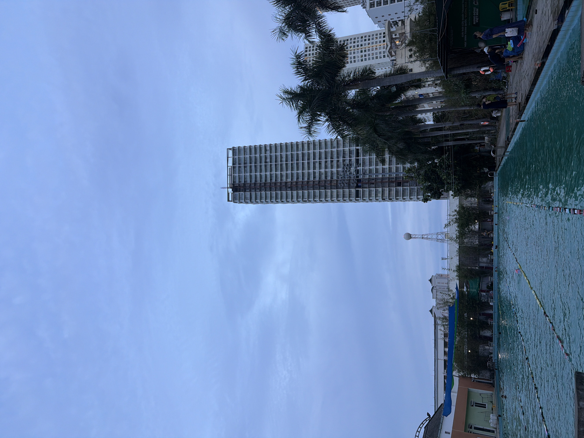 The image depicts an outdoor swimming pool facility in Nha Trang, Vietnam. The pool is large, with lane markers visible, and several people are swimming. The surrounding area includes palm trees, some buildings, and a tall, modern high-rise building that appears to be under construction. The sky is overcast. On the right side of the image, there are several people gathered near a covered area, with some objects on the ground that could be bags or equipment. A sign is visible behind them, though the text is not legible. The overall impression is of a public recreational space in an urban setting.