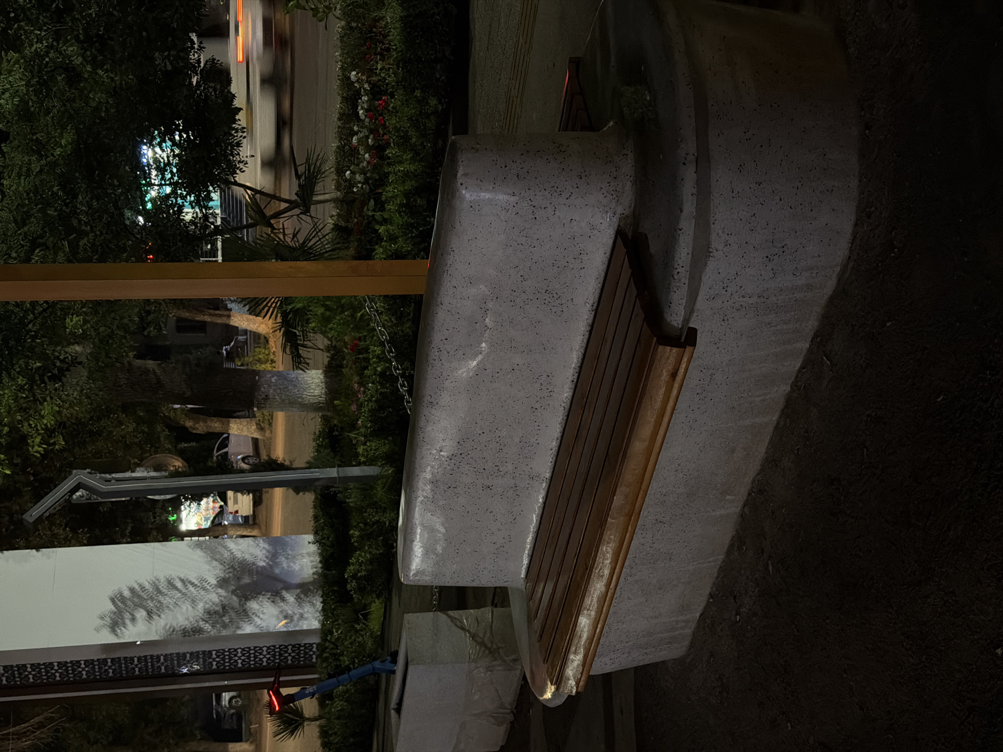 A concrete park bench with wooden slats for seating is positioned in the foreground, to the right of a concrete planter box. Behind the bench, a street is visible with blurry car lights indicating movement. Trees and bushes line the street, and in the background, a building with illuminated signage can be seen. The scene appears to be a public space in Chirchik, Uzbekistan, at night.