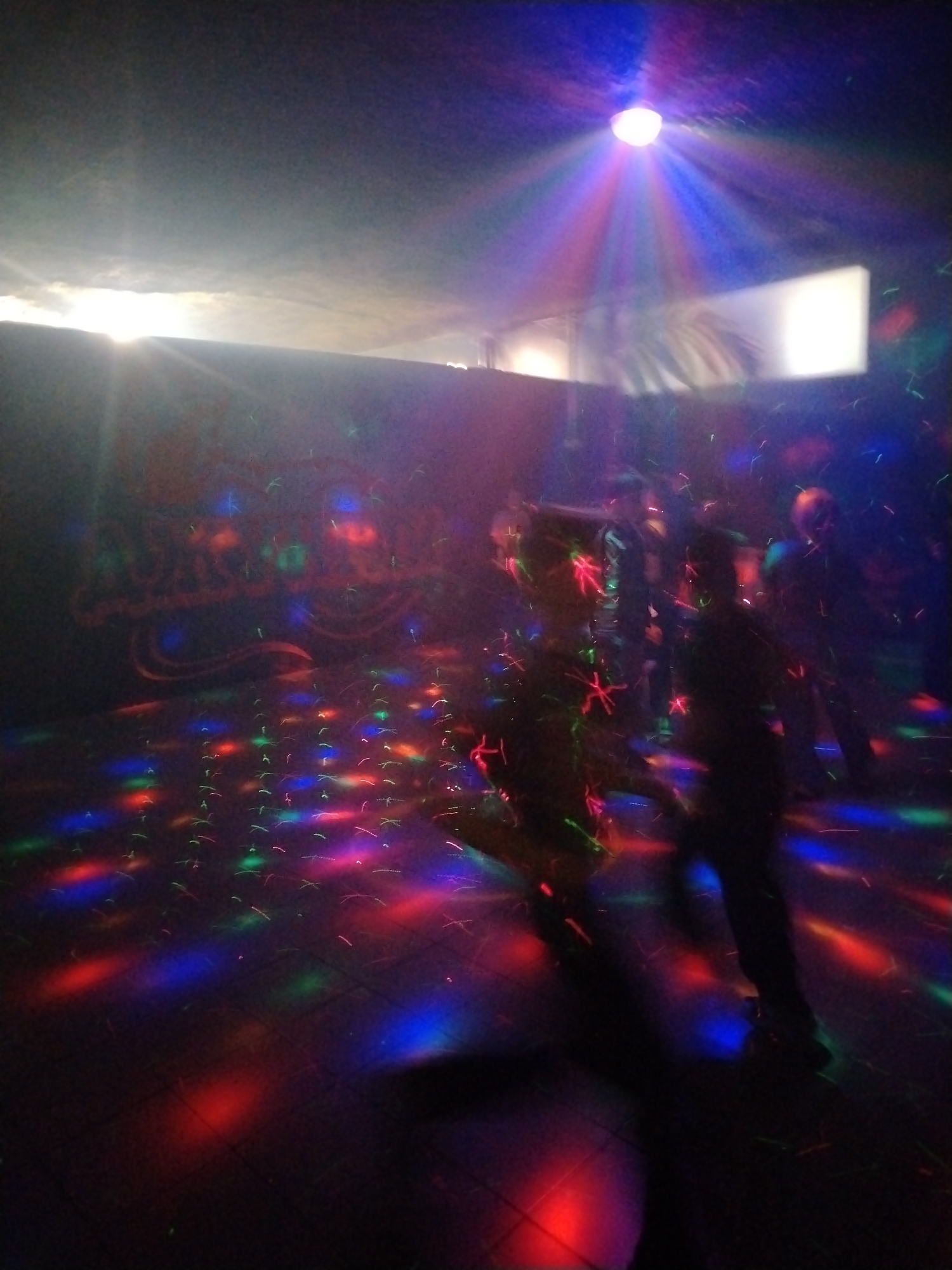 The image shows an interior scene, likely a nightclub or event space, in Заволжье, Russia. The space is dark with colored lights projected on the floor and walls, creating a disco-like effect. There are several people present, their forms blurred due to motion, suggesting they are dancing. A single spotlight is directed downward from the ceiling, casting a blue and red hue. A bright, unidentified light source is visible through a window or opening in the upper left corner of the frame. A white rectangular object is seen in the upper right, likely a screen or blank wall, also illuminated. The floor appears to be tiled.