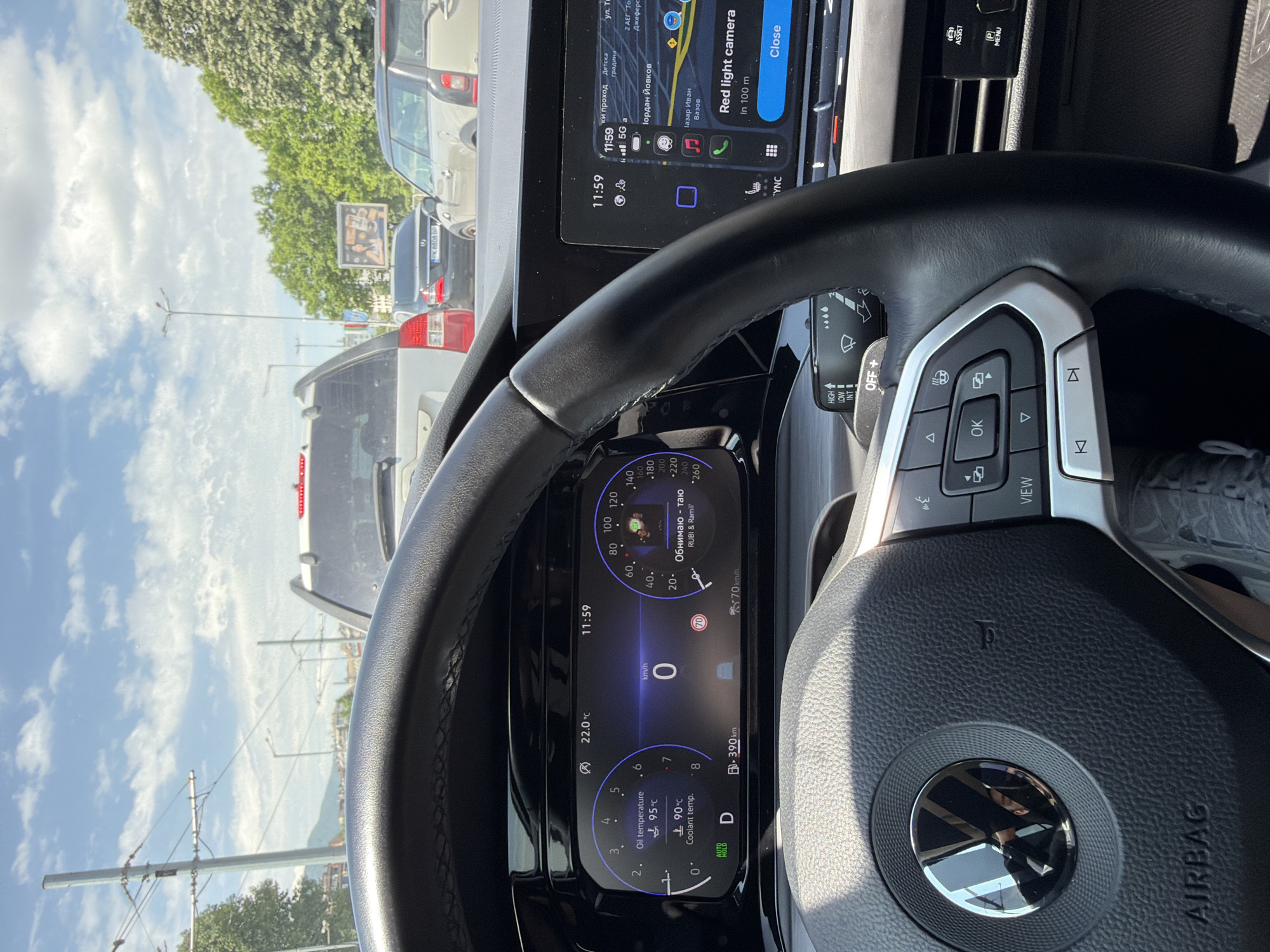 The image shows the interior of a Volkswagen car. The dashboard displays a digital instrument cluster indicating 0 km/h, oil temperature at 95°C, coolant temperature at 90°C, and a range of 390 km. The infotainment system shows a navigation app with a "Red light camera in 100m" alert. The location is inferred to be in Sofia, Bulgaria, based on the street names "Тордан Йовков" and "Газар Иван Вазов" displayed on the navigation screen. The screen also displays "PK 8808 BP" on a vehicle that can be seen through the front windshield. A billboard is visible in the background, further confirming it is a street scene in the city. The car is stationary, in drive mode (D) with automatic brake hold "AUTO HOLD" enabled, as indicated on the dashboard. The steering wheel is in view with control buttons.