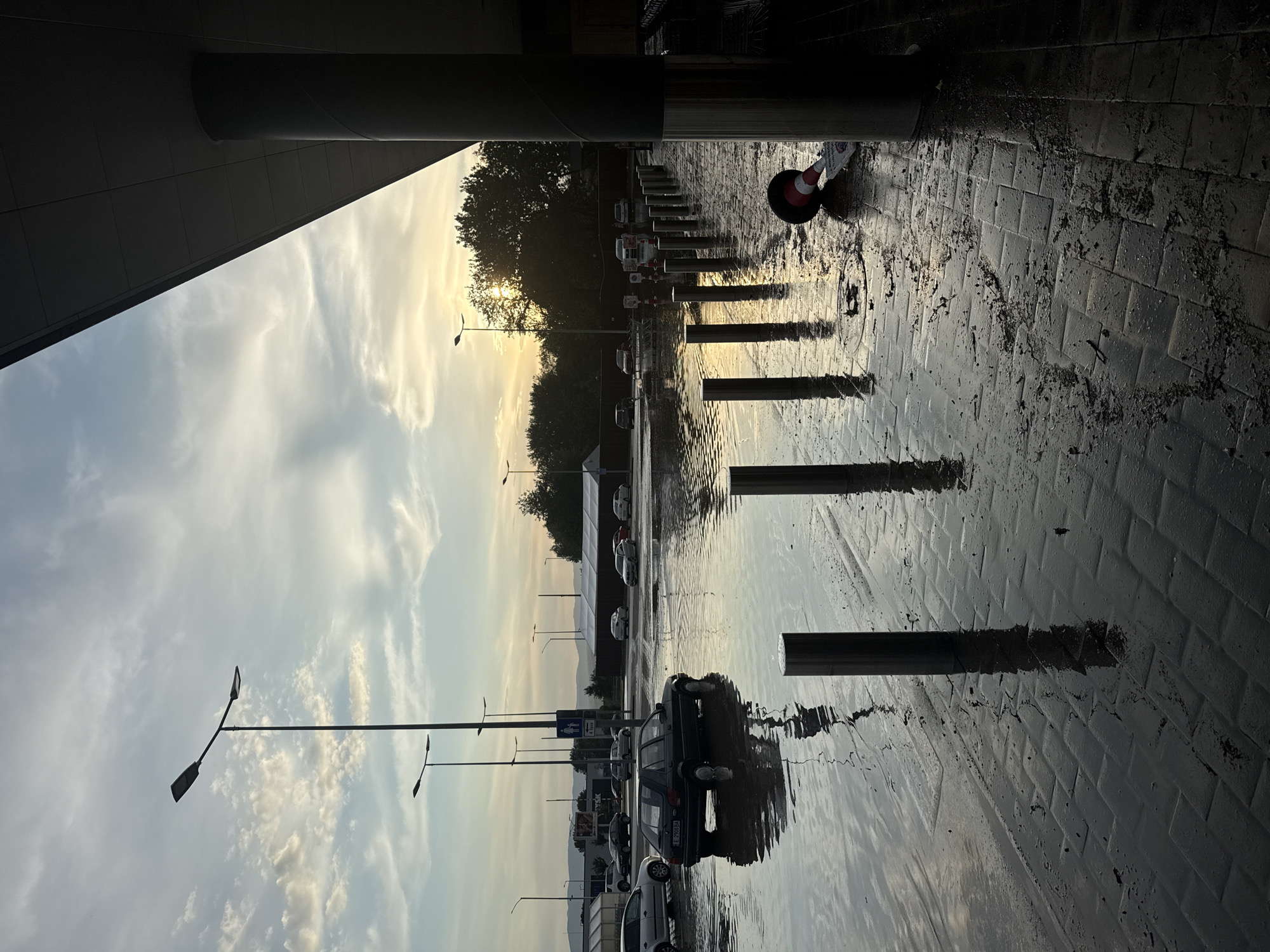The image depicts a wet outdoor scene, likely a parking area or service road next to a building, under a sky suggesting either dawn or dusk. The dominant feature is the reflective, puddle-laden ground, indicating recent rainfall.
On the left, a large, dark architectural structure, possibly an overhang or part of a building, casts a deep shadow. A substantial cylindrical column supports this structure. To the right of the building, a paved area, possibly made of dark tiles or cobblestones, extends into the mid-ground. This surface is covered in numerous puddles, creating strong reflections of the sky, distant objects, and the warm light of the setting or rising sun.
A line of reflective bollards stretches across the mid-ground, their silver surfaces and shadows clearly mirrored in the water. Further right, a red and white traffic cone lies on its side in a puddle. In the mid-distance, several cars are parked, their shapes and reflections visible. One dark-colored station wagon or hatchback is prominent, with its license plate "P6239BA" partially legible. Behind the parked cars, a dense line of trees is silhouetted against the bright sky, with the sun's warm glow peeking through the foliage. In the far background, hazy mountains outline the horizon.
Tall lampposts with horizontal arms are scattered across the scene, their silhouettes reaching into the sky. The sky itself is filled with scattered clouds, displaying a gradient from a warm yellow-orange near the horizon to lighter blues and whites higher up. No people are visible.
The scene conveys a quiet, post-rain atmosphere, with the soft light and extensive reflections creating a serene mood. The location context is Blagoevgrad, Bulgaria.
