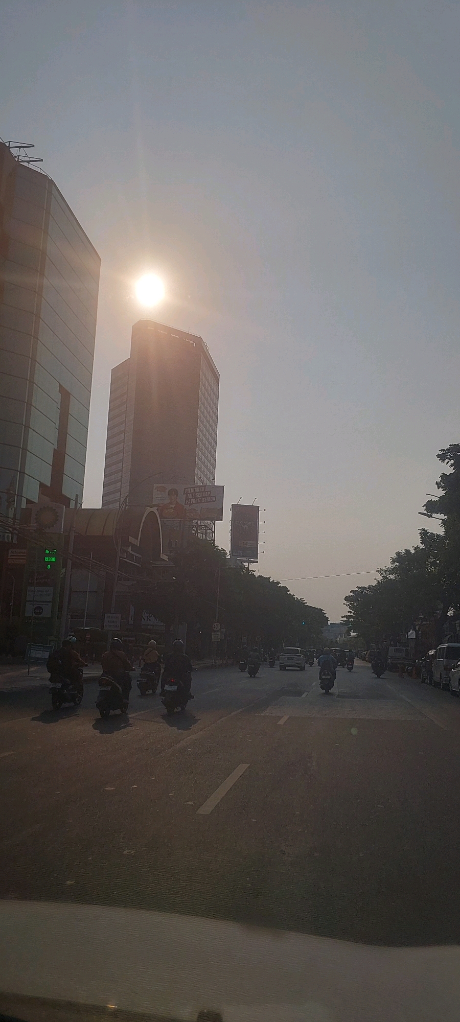 This image depicts a street scene in Surabaya, Indonesia, under a bright, hazy sun. Two modern high-rise buildings dominate the background, one with a glass facade on the left and another taller, darker building in the center. The sun is partially obscured by the buildings, creating a lens flare effect.
The street is a multi-lane road with active traffic. Numerous motorcycles are the primary mode of transportation visible, with riders wearing helmets and casual clothing. A few cars and other vehicles are also present. The road surface appears to be asphalt, with clear lane markings.
Along the sides of the street, there are trees, streetlights, and billboards. One billboard displays an advertisement with text in Indonesian. A traffic light can be seen, currently showing a green light. The overall atmosphere suggests a busy urban environment during daylight hours, possibly late afternoon given the angle of the sun. The haze in the sky may indicate air pollution or atmospheric conditions common in tropical climates. The perspective is from inside a vehicle, looking out through a windshield, suggesting the photo was taken by someone in transit.