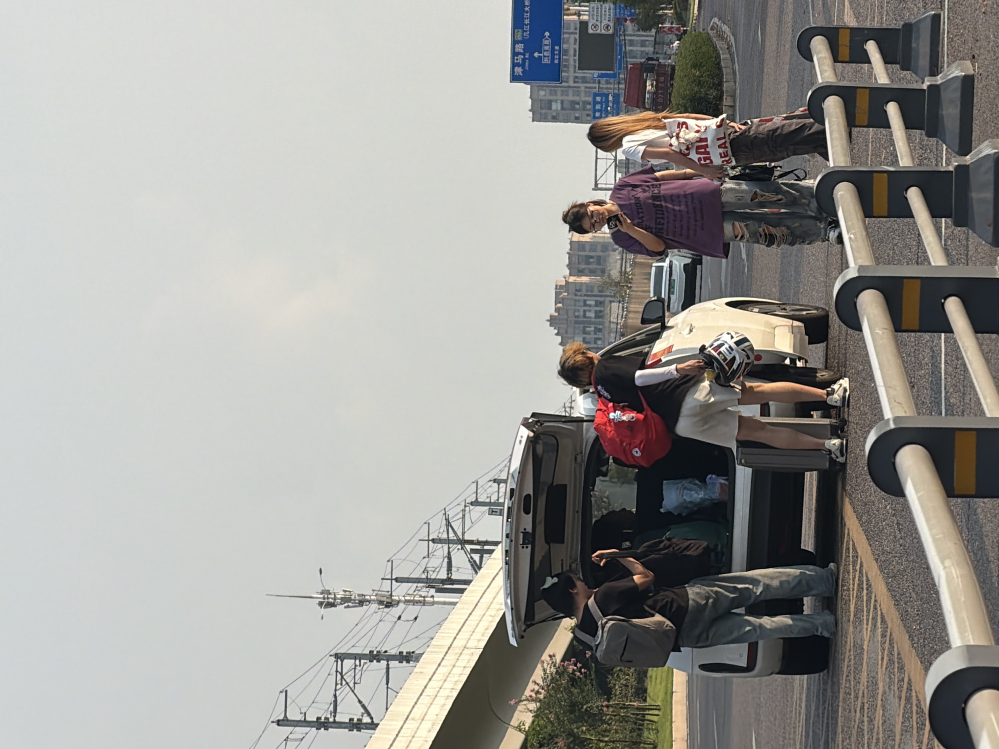 Four individuals are gathered around the open trunk of a white SUV on a city street. Two individuals are on the left, one wearing a black shirt and backpack, and another partially obscured behind the SUV. On the right, one individual in a purple t-shirt and ripped jeans is looking at their phone, while another in a white t-shirt and patterned pants stands beside them. Luggage, including a red backpack and a suitcase, is visible. The background features a highway overpass with electrical wires, buildings, and traffic signs. One sign is in Chinese and indicates "Jingma Road" and "Jiangjin Yangtze River Bridge". The overall environment suggests an urban setting in Jiangjin, China.