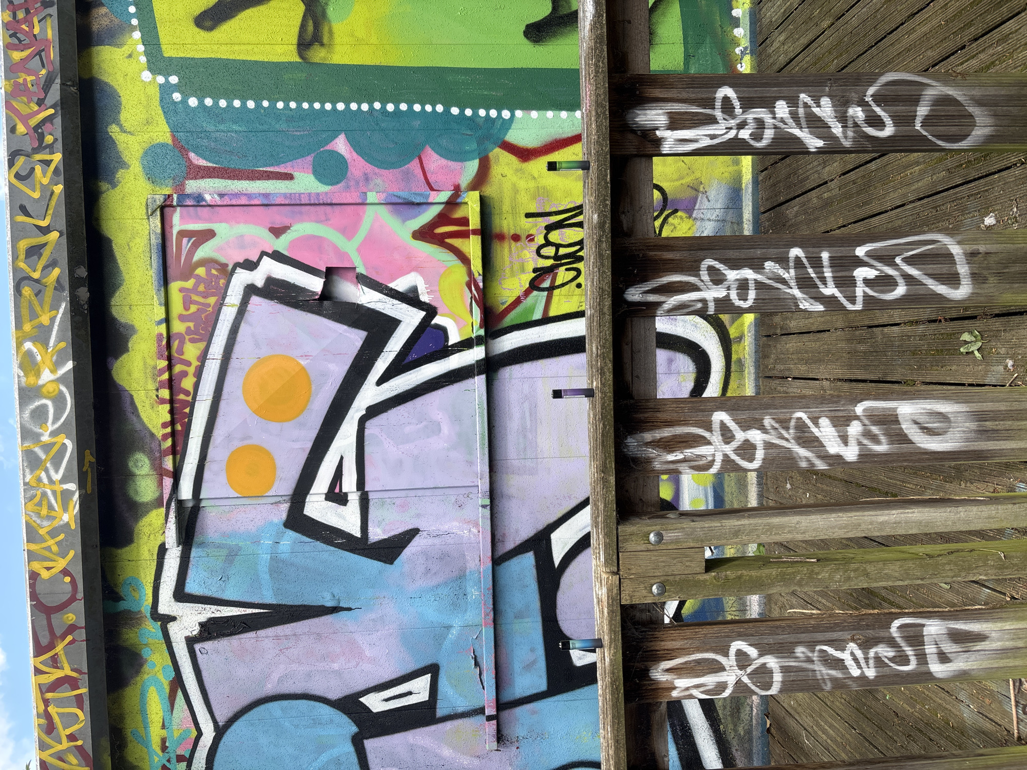 The image shows a concrete wall covered in extensive graffiti in various colors, including green, yellow, pink, purple, and blue. A wooden structure, possibly part of a staircase or walkway, stands against the wall. White graffiti is spray-painted on the wood. The scene could be located in an urban environment, potentially in London, given the prevalence of graffiti in certain areas of the city.