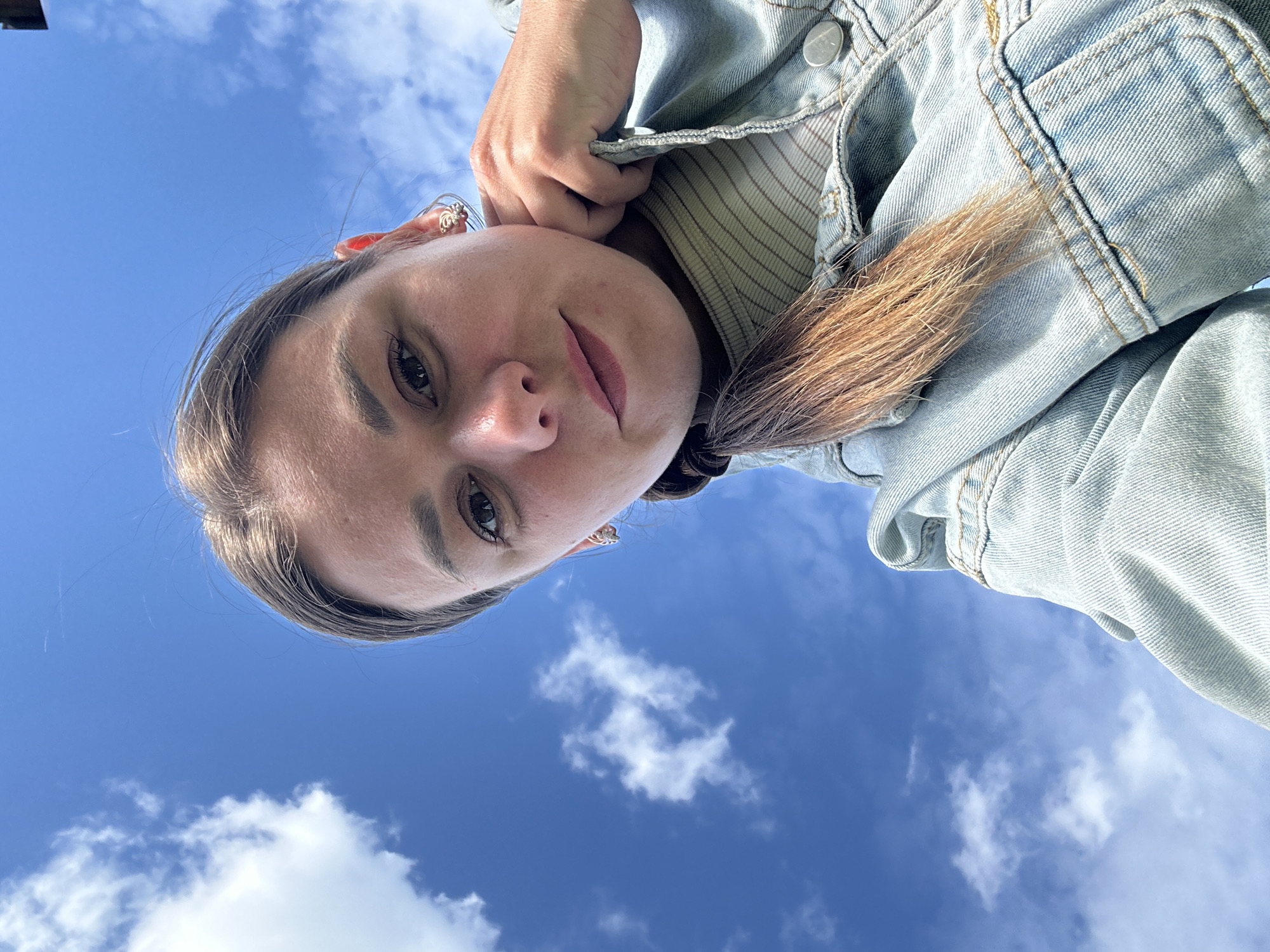 The media file is a close-up, low-angle selfie of a young woman against a bright blue sky with scattered white clouds. She is wearing a light blue denim jacket over a striped shirt. Her hair, which is long and brown, is falling forward, obscuring part of her face and clothing. She is looking directly at the camera with a slight smile. The lighting suggests it is daytime, likely late morning or afternoon, with sunny weather. The image is taken outdoors, with the vast sky as the background. There are no visible text elements in the media file. The subject's pose and the angle of the shot create a sense of looking up at her against the expansive sky.