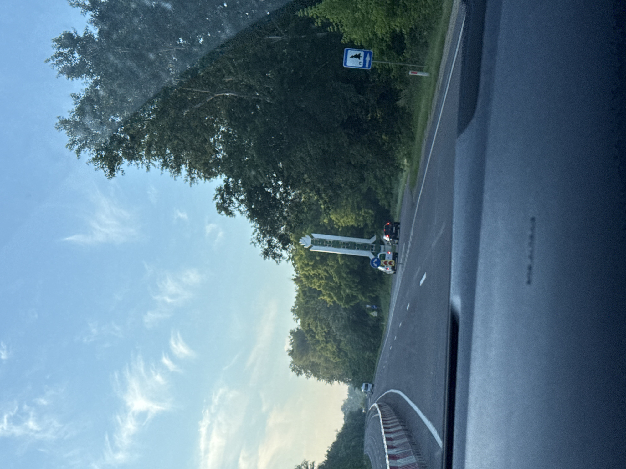 The image depicts a view from a vehicle on a highway, likely in or near the city of Cojușna, Moldova. The road is paved, with white lane markings and a concrete barrier with red and white reflective markings on the left side, suggesting ongoing construction or maintenance. Several vehicles are visible on the road, including a white van further ahead and two cars closer to the foreground on the right lane.
On the right side of the highway, there is a tall, white, monument-like structure with green accents. Adjacent to it, a blue and white road sign with a silhouette of a tree and an arrow pointing to the right is visible. The surrounding environment is lush and green, dominated by dense trees and foliage. The sky is a clear blue with wispy white clouds. The image is taken during daylight, possibly late afternoon or early evening, given the soft lighting. The foreground shows the dashboard of the vehicle from which the photo is taken.