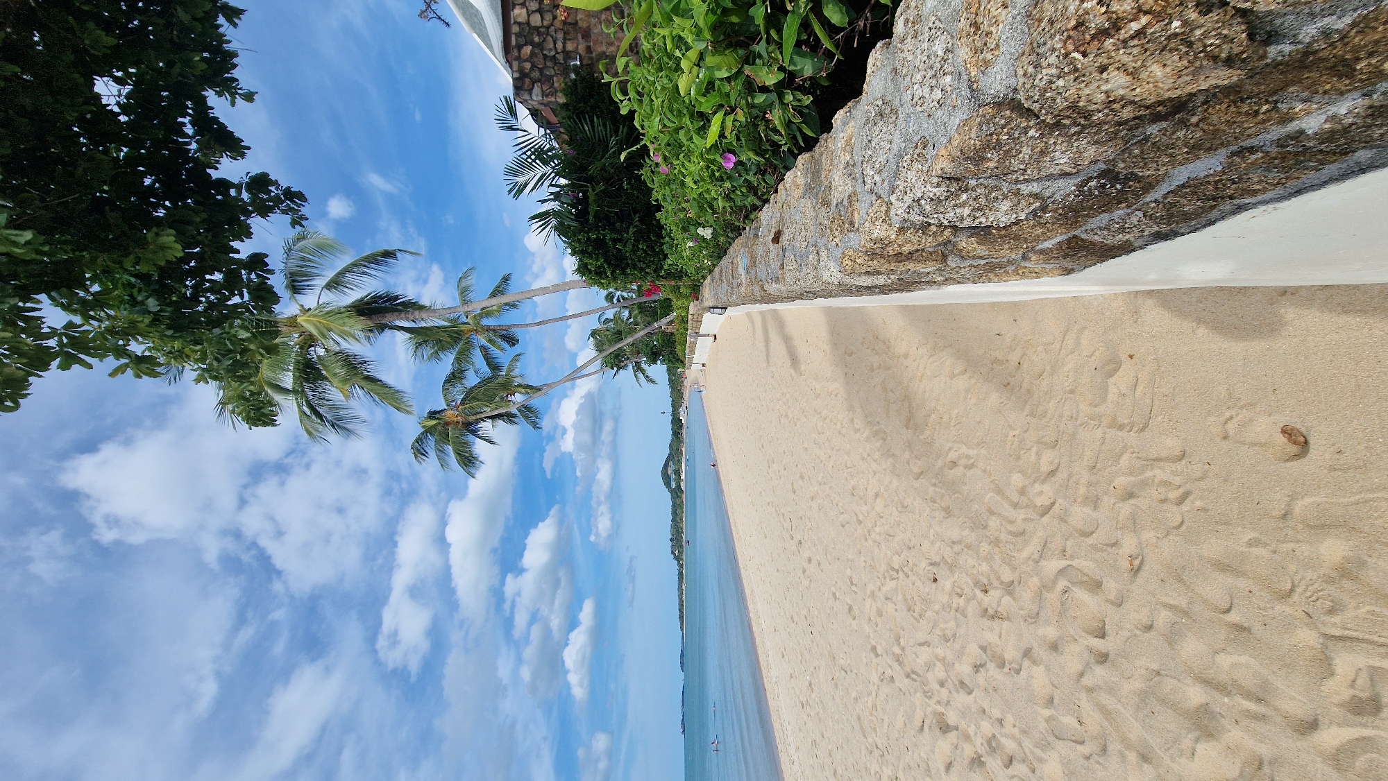 The image captures a sunlit tropical beach scene in Baan Mae Nam, Thailand, characterized by a bright blue sky with scattered white cumulus clouds. The foreground features a wide expanse of light beige sand, imprinted with numerous footprints and showing minor undulations. A small, dry brown leaf rests on the sand in the lower right.
A low stone wall, composed of irregular, rough-hewn reddish-brown and gray stones, with a distinct white painted base, extends diagonally from the lower right corner towards the mid-ground on the right side. Behind this wall, dense green foliage and shrubbery are visible, interspersed with small pink and white flowers. Further back on the right, partially visible is a building with a textured stone exterior and a light-colored roof, surrounded by more lush greenery.
Several tall, slender palm trees with green fronds lean slightly over the beach and wall from the right side of the image. A canopy of broader, dark green leaves from another tree hangs into the frame from the top right.
To the left, the calm, light blue ocean stretches towards the horizon. Several small, indistinct human figures are visible in the shallow water, appearing to be swimming or wading. A few additional figures are sparsely distributed on the distant beach. Along the far coastline, a hazy landmass or hills are visible under the sky. The scene is brightly illuminated by natural light, casting distinct shadows from the palm trees onto the sand and highlighting textures throughout the environment.