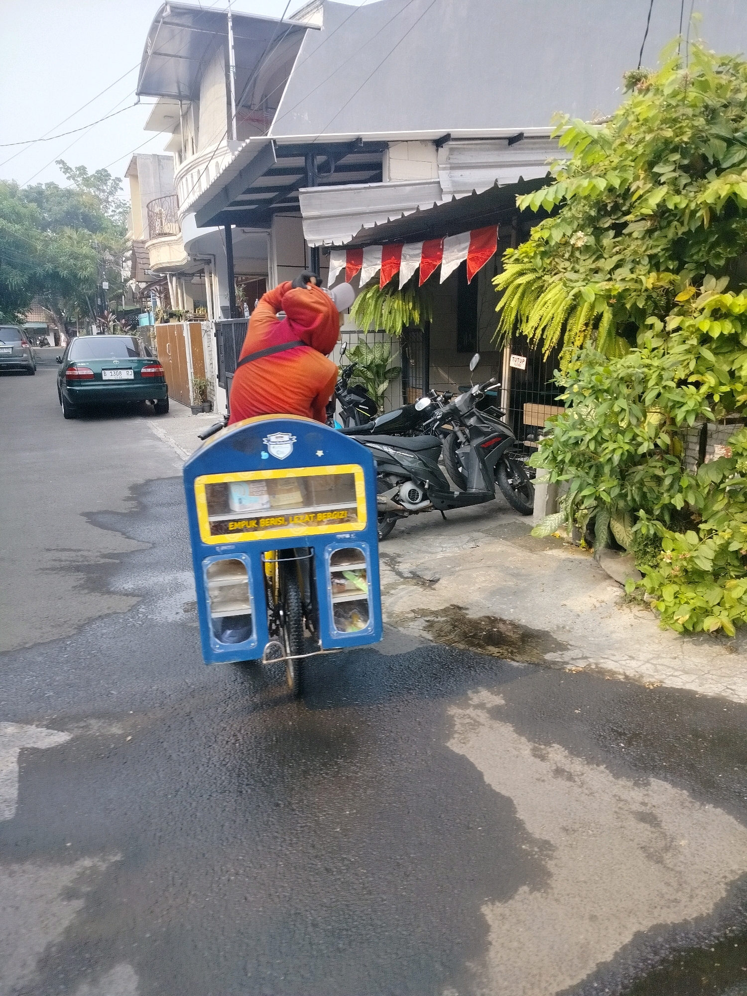 An outdoor, eye-level, slightly angled, full shot captures a street scene in East Jakarta, Indonesia, on a sunny day. A person wearing an orange hooded jacket is riding a blue and yellow three-wheeled bicycle with a cargo box. The cargo box is adorned with a logo and text that reads "EMPUK BERISI, LEZAT BERGIZI!". The bicycle is on a wet asphalt road. To the right, there are motorcycles parked on a concrete sidewalk. Behind the motorcycles, lush green trees and plants are visible, bordering a building with red and white decorative flags. In the background, a dark green car is parked on the side of the road. The street is lined with buildings and trees, suggesting a residential or commercial area.
