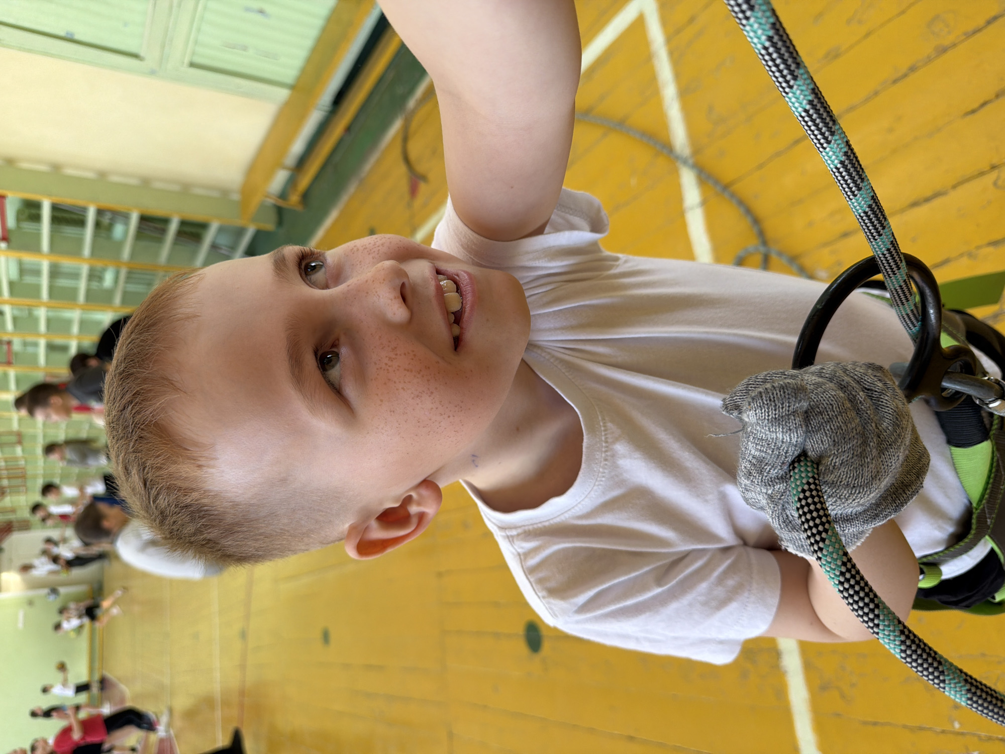 A fair-skinned, freckled boy in a white t-shirt is suspended in the air with a climbing harness and rope in what appears to be a gymnasium. He is wearing a grey glove and gripping the rope. In the background, other people are visible on the gymnasium floor. The environment suggests an indoor athletic facility with a yellow floor. The specific location, Izhevsk, Russia, is not identifiable from the visual information.