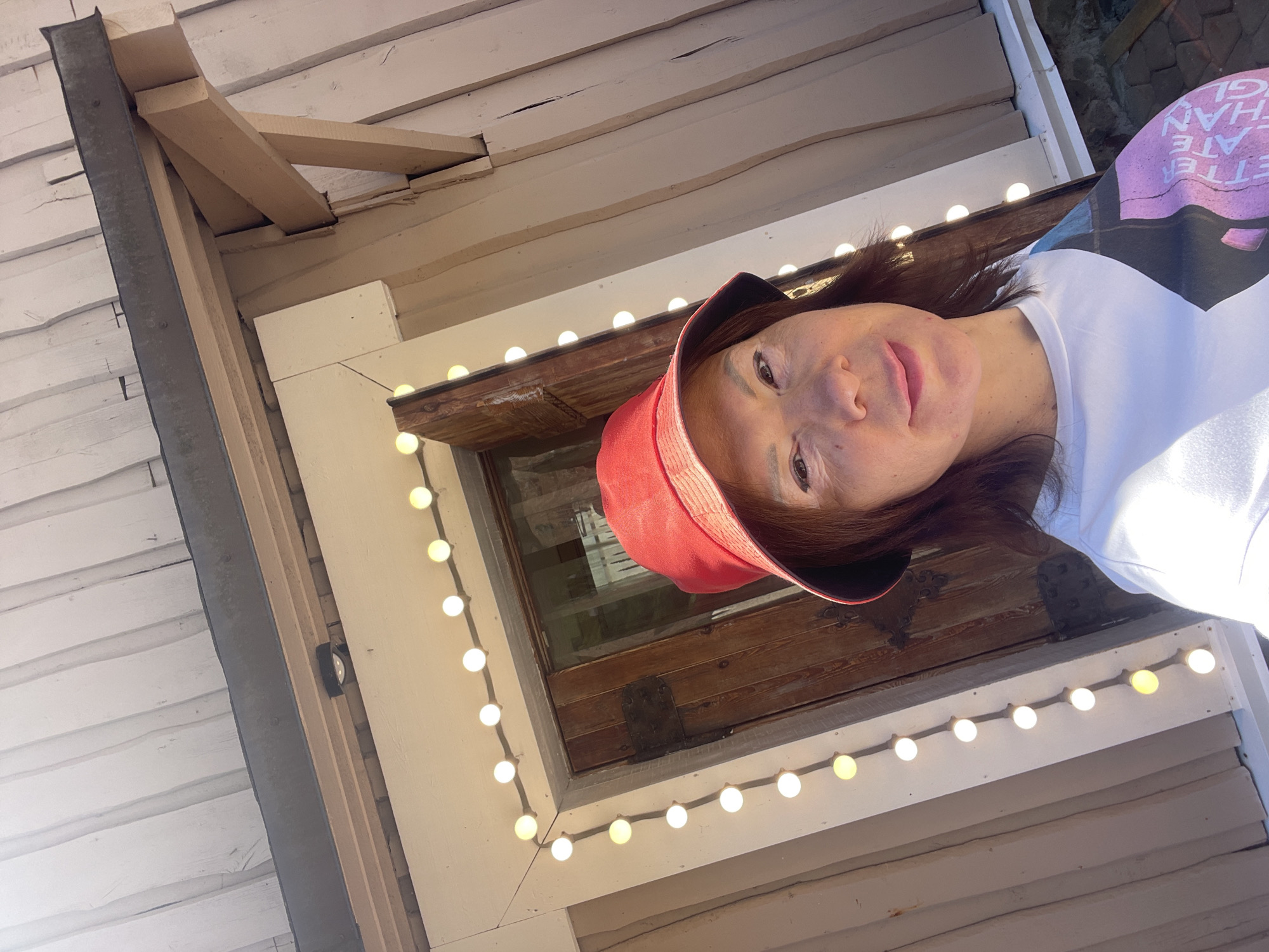 A woman wearing a red bucket hat and a white t-shirt stands in front of a wooden door. She is taking a selfie, and the photo is angled from below. The door is decorated with a string of white lights. The building appears to be made of wood with a weathered texture. The lighting suggests it is daytime, and the sun is casting shadows. The woman's expression is neutral, and she is looking at the camera. The image is an outdoor scene. The provided crops highlight details of the building's construction and the string lights.