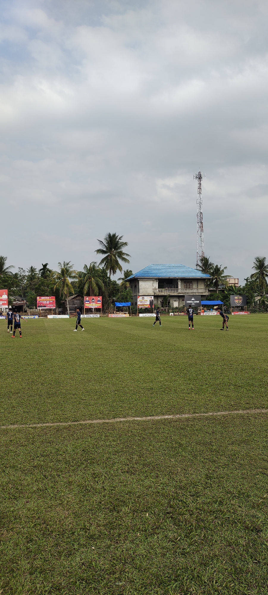In Kilometer Delapan, Indonesia, a soccer game is underway on a bright, overcast day. The playing field is a lush green expanse marked with white lines, a single white line stretching across the lower portion of the frame. Several players, dressed in dark uniforms with white accents, are scattered across the field, some in motion, others standing. A few individuals are gathered near the sidelines, close to a row of advertising banners. Behind the field, a two-story building with a bright blue roof stands prominently, flanked by palm trees and other vegetation. Further in the background, a tall, multi-tiered communication tower pierces the sky, its lattice structure visible against the cloudy atmosphere. The overall scene depicts a typical local sporting event in a semi-rural Indonesian setting.