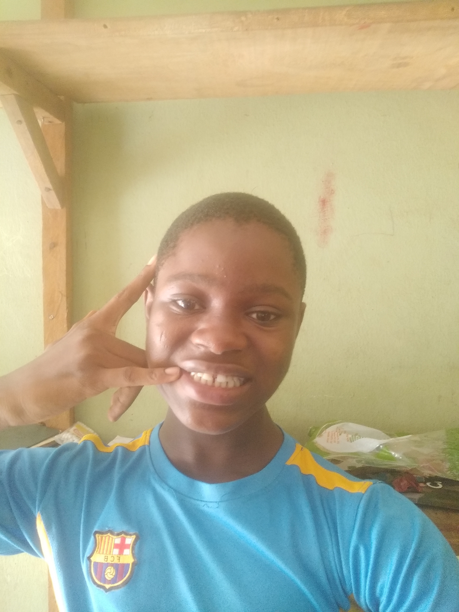 A close-up, brightly lit portrait features a young person with dark skin and short hair, smiling directly at the camera. They are making a "call me" or "shaka" hand gesture with their right hand, positioned near their cheek, conveying a cheerful and playful mood. The individual wears a light blue short-sleeved t-shirt with distinctive yellow, wavy accents on the shoulders. Prominently displayed on the left chest is a crest-shaped logo, likely representing FC Barcelona, with the letters "FCB" and a football motif beneath striped patterns. The background is a plain, light green wall. Wooden shelves or supports are visible in the upper left and top of the frame, including a diagonal wooden brace. A faint, reddish vertical smudge is present on the wall to the right of the person's head. Blurry objects, possibly white plastic bags, are visible behind the person's right shoulder. The soft, natural lighting suggests an indoor setting during daylight hours.