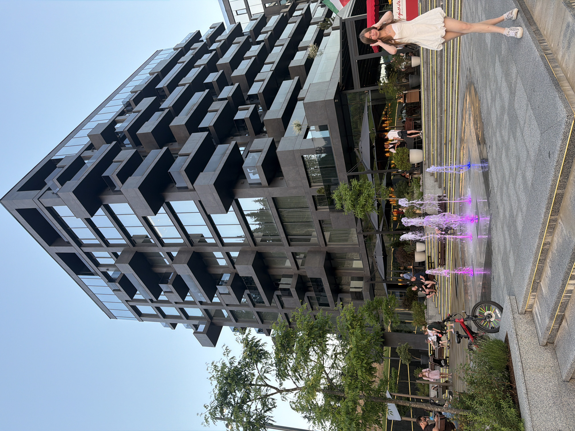 This image depicts an outdoor scene in Polianytsia, Ukraine, featuring a modern, multi-story building with a dark, textured facade and numerous glass-enclosed balconies. In the foreground, a young woman in a white dress and sneakers stands with her hands on her head, facing the camera. To her left, a fountain with colorful illuminated water jets is active. A red bicycle with multicolored spokes is parked near the fountain. Several people are seated on benches and at tables in the background, with some dining outdoors under umbrellas in front of the building. Lush green trees and foliage are visible on the left side of the image. The sky is clear and blue, indicating daytime.