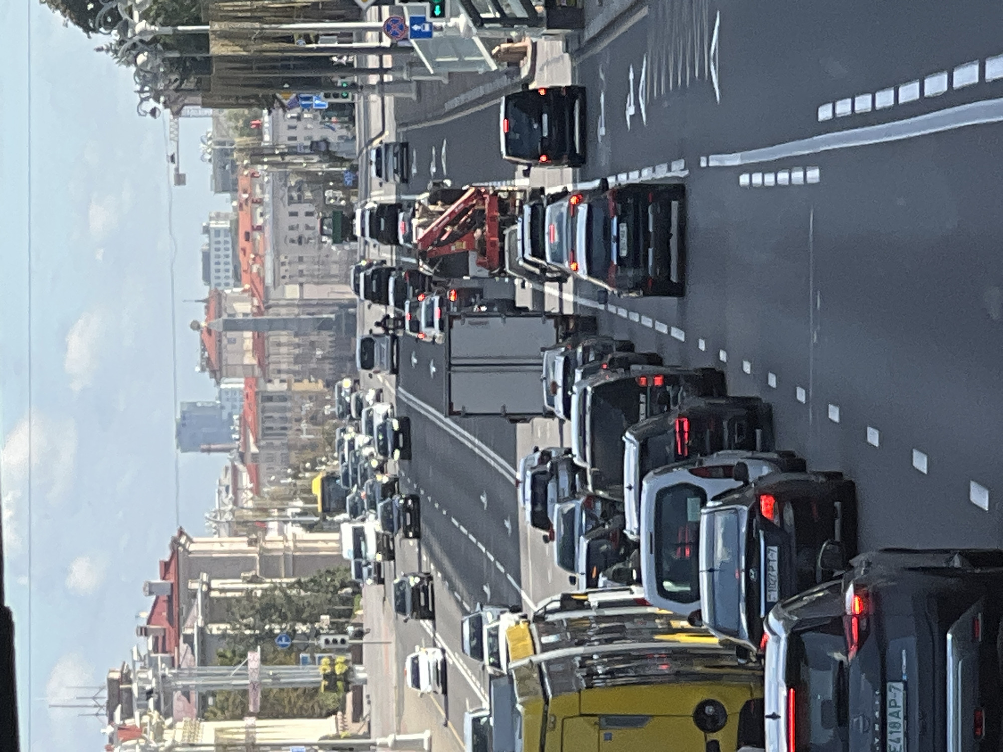 The image depicts a congested traffic scene on a wide, multi-lane avenue in Minsk, Belarus. The street is lined with classical European-style buildings featuring red-tiled roofs and ornate architecture. The sky is partly cloudy.
Vehicles are bumper-to-bumper, indicating a significant traffic jam. Several types of vehicles are visible, including passenger cars of various makes and models, a yellow bus or trolleybus on the left, a white van, and a truck with a red crane. The traffic is flowing in both directions.
Street signs are visible, including a blue traffic sign with a white arrow and a red circle with a diagonal bar, and another blue sign with a white pedestrian symbol. Traffic lights are also present. The road is marked with white dashed lines indicating lanes and arrows for directional traffic.
In the distance, taller modern buildings are visible, suggesting a mix of old and new architecture in the city. The overall impression is of a busy urban environment experiencing a traffic standstill.