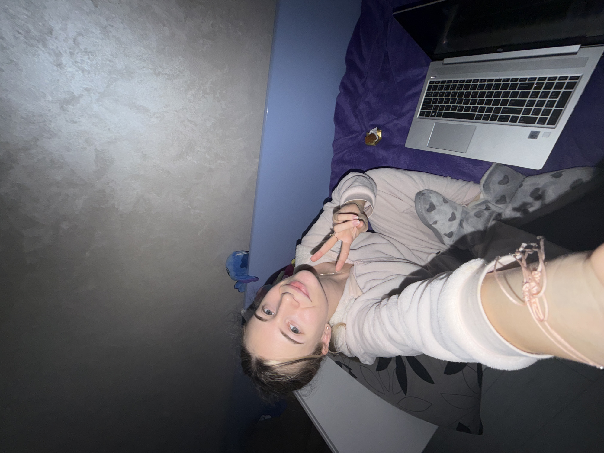 A young fair-skinned woman lies on a dark purple plush bed with gray slippers that have white hearts on them. She wears a white terry cloth robe. She is making a peace sign with her left hand. She is taking a selfie with her right arm extended. She has a pale pink string bracelet on her right wrist. A silver laptop computer is open on the bed near her, with a piece of candy beside it. A blue stuffed animal is on the headboard of the bed. There is a gray wall on the left. The location may not be in Plovdiv, Bulgaria.
