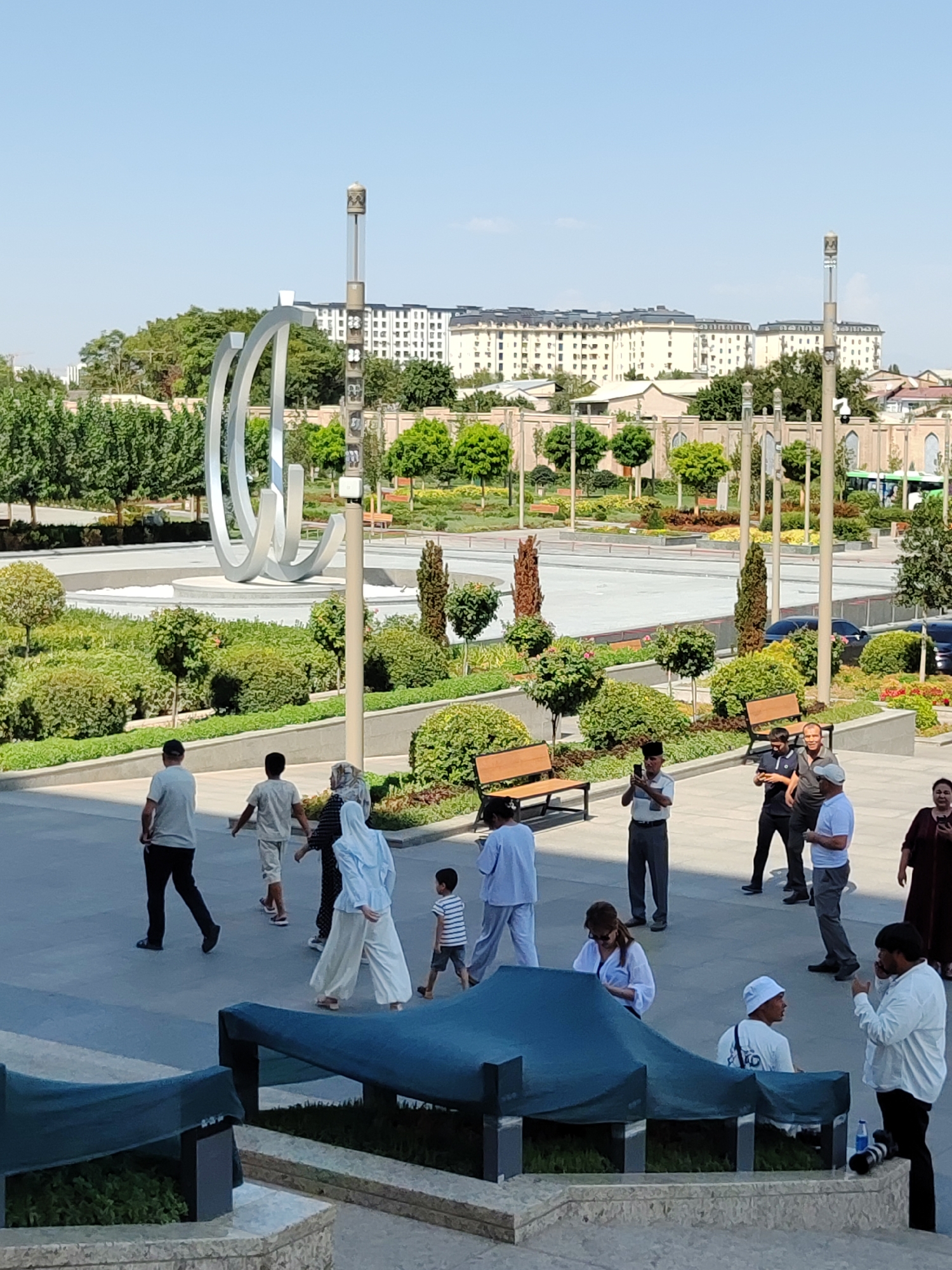 This is a street-level, full-frame, eye-level, medium shot capturing a sunny outdoor scene in Tashkent, Uzbekistan, likely in a public park or plaza. The foreground features a modern architectural element covered by a dark blue tarp, with greenery visible beneath. Several individuals are scattered throughout the midground, engaged in various activities.
To the left, a man in a grey t-shirt and dark pants walks away from the camera. Next to him, a young person in a light grey t-shirt and light pants walks in the same direction. A woman in a light blue hijab and light pants is walking towards the right, and a young child in a striped t-shirt and shorts walks beside her. Further back, a man in a white t-shirt and dark pants is taking a photo with his phone. Two men in dark shirts stand nearby, one looking at his phone and the other engaged in conversation. A woman in a dark maroon dress stands to the right, looking away from the camera.
In the background, a large, abstract silver sculpture stands prominently in a circular fountain area. Lush green trees and manicured bushes surround the sculpture. Behind the park, a multi-story building with a light facade and dark roofs dominates the skyline, suggesting an urban environment. The sky is clear and blue. The overall atmosphere appears to be one of leisure and casual activity on a bright, warm day.