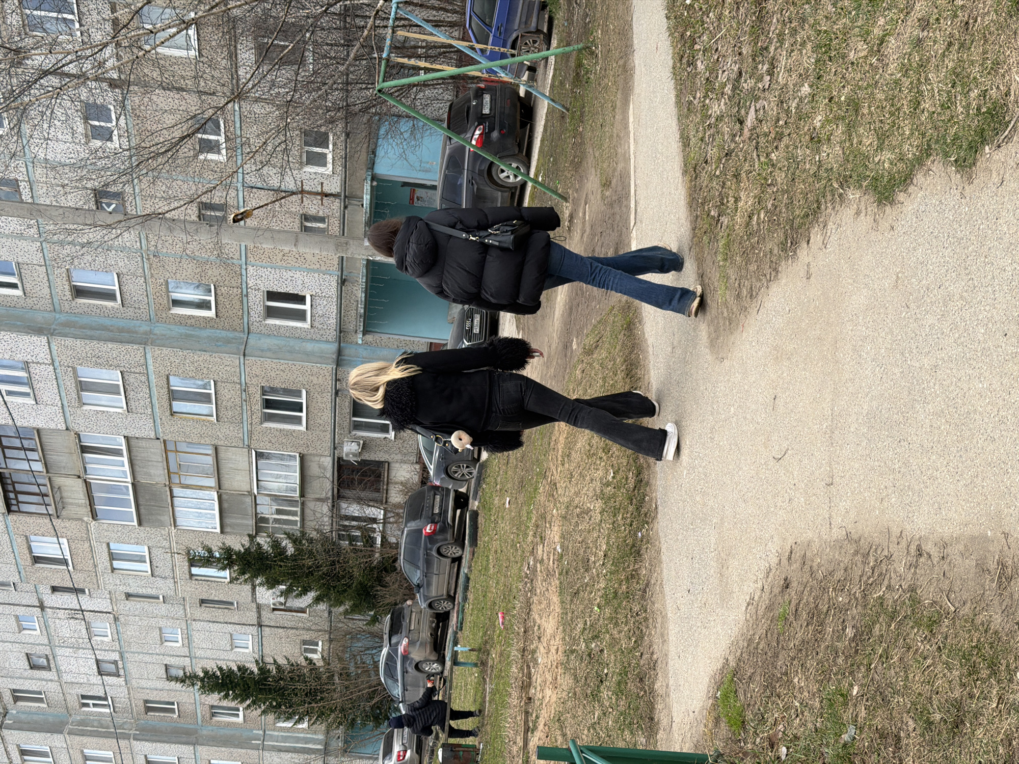 Two women are walking on a sidewalk. The woman in the front has long blonde hair and is wearing a black jacket with fur trim, black jeans, and white sneakers. The woman behind her has dark hair and is wearing a black puffer jacket, blue jeans, and brown flats. They are walking away from the viewer.
To the left of the sidewalk, there is a grassy area with several parked cars. Behind the cars is a large, gray apartment building. In the background, there is a swing set. The photograph appears to be taken in Perm, Russia, based on the architectural style of the apartment building and the makes of the cars.