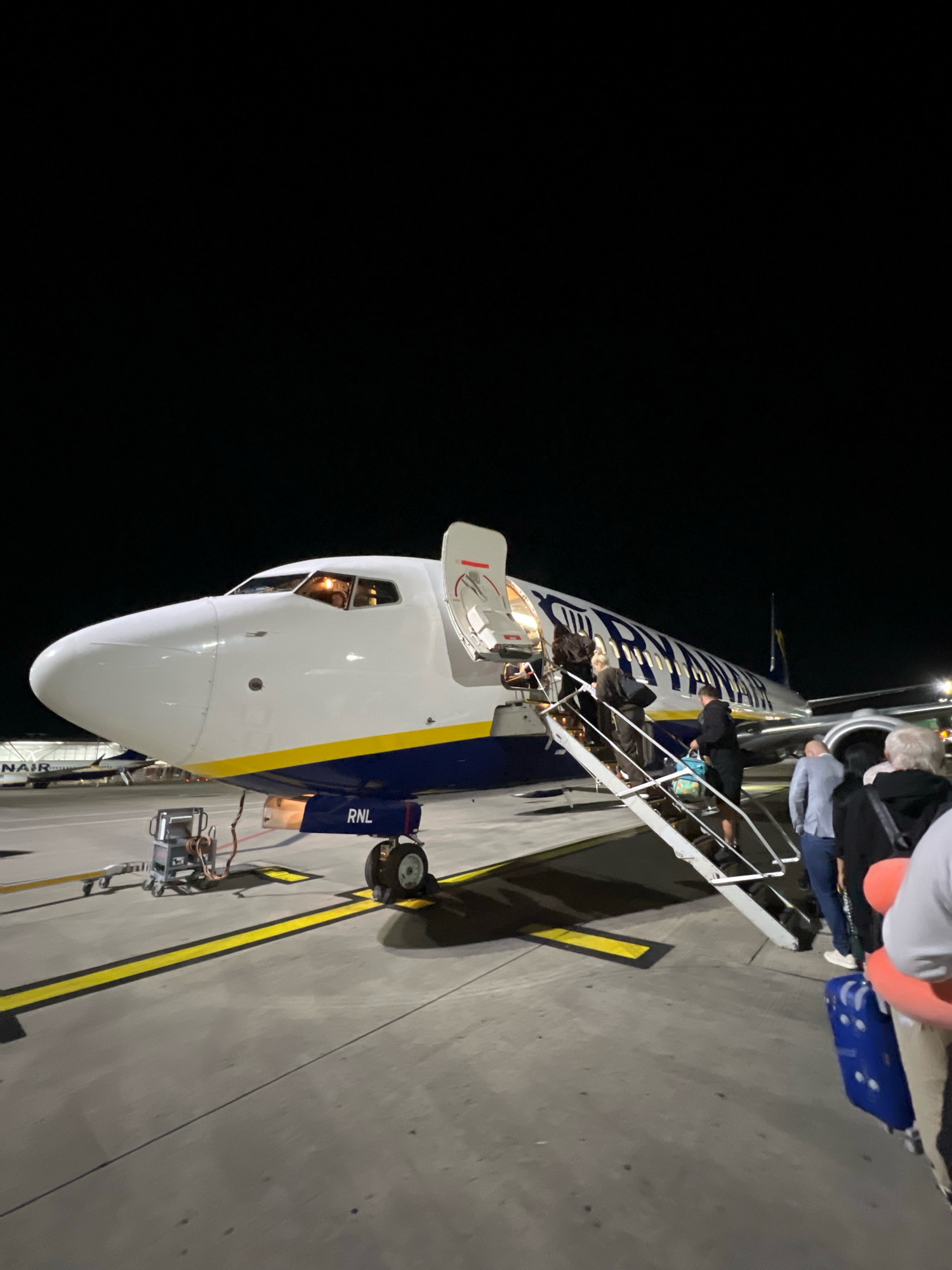 An exterior, eye-level shot captures a Ryanair airplane at night on an airport tarmac, likely at London Stansted Airport in Uttlesford, United Kingdom, as suggested by the airline and airport signage. The airplane, identified by the registration "RNL" near the front landing gear, has its forward cabin door open, with a set of mobile stairs leading up to it. Several passengers are in the process of boarding, ascending the stairs with luggage. A blue and yellow livery adorns the aircraft. In the background, another aircraft with "RYANAIR" branding is visible. The tarmac is marked with yellow lines. The scene is illuminated by artificial lighting, casting shadows on the ground.