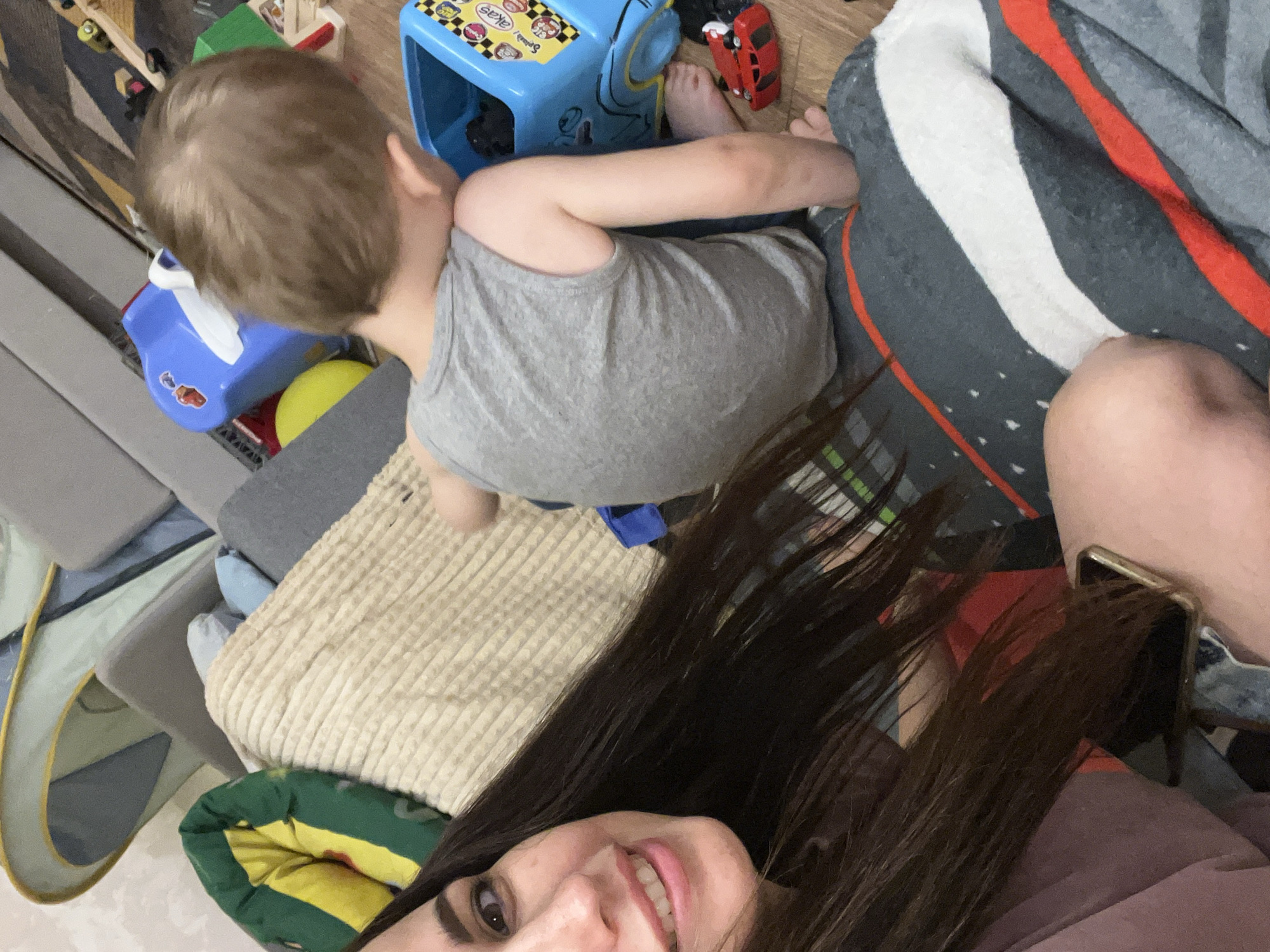 A medium shot shows a woman and a young child on a floor covered with a blanket. The woman is on the left, with her face turned towards the camera, smiling. She has long, dark hair and is wearing a pink top. The child is on the right, facing away from the camera, playing with toys. He is wearing a grey tank top and grey shorts. The floor is cluttered with children's toys, including a blue toy truck, a red toy car, and a yellow ball. In the background, there are several large cushions and a tent-like structure. The scene appears to be a living room or playroom.