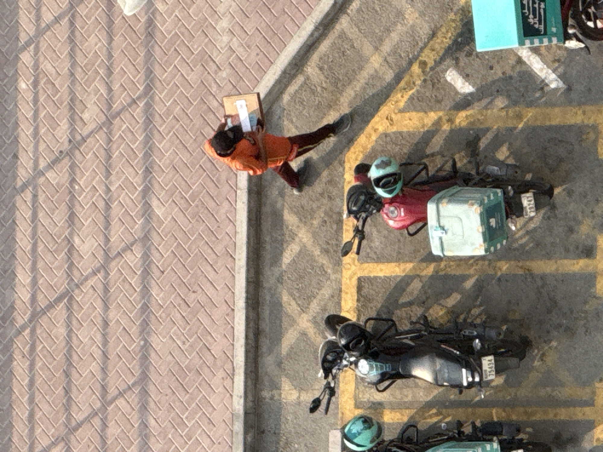 An aerial shot captures a delivery person walking from right to left, carrying a flat cardboard box. The individual is wearing an orange hooded jacket and dark pants with a yellow stripe. They are on a paved surface with a herringbone brick pattern in the upper portion of the frame, which transitions to asphalt with yellow parking lines.
In the lower half of the image, three motorcycles are parked in tandem. The first motorcycle, on the left, is partially visible and has a teal helmet resting on its handlebars. The second motorcycle is black with a teal helmet. The third, a red motorcycle, has a light blue cargo box on the back and also features a teal helmet. The license plate of the black motorcycle reads "75434," and the red motorcycle's license plate reads "29247."
The scene appears to be an outdoor parking area in Dubai, United Arab Emirates, possibly near a residential or commercial building, given the arrangement of parked delivery vehicles. The lighting suggests it is daytime, with distinct shadows cast by the motorcycles and potentially an overhead structure.
