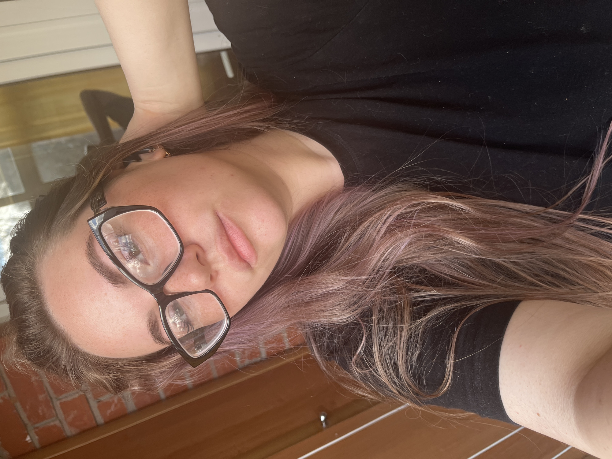 The image shows a young woman lying down and looking towards the camera, positioned with her head tilted. She has long, wavy hair with distinct streaks of brown, blonde, and muted pink or lavender tones. She is wearing large, black-framed glasses that cover a significant portion of her face. Her complexion is fair, with visible pores and a few minor blemishes. Her lips are a natural light pink, and her eyebrows are dark and well-defined.
She is dressed in a plain black short-sleeved t-shirt, on which some small specks of lint or dust are visible. One arm is visible in the upper part of the frame, bent at the elbow, suggesting it might be resting behind her head or neck.
The background consists of several elements, suggesting an indoor or semi-outdoor setting. In the upper left, there is a light-colored, possibly white, horizontal surface. Below this, and extending to the bottom right, is a warm-toned wooden surface, possibly flooring or paneling. A section of reddish-brown brickwork is visible in the lower left corner. There's also a reflection on a flat surface behind her arm, showing a distorted dark shape, possibly a person or furniture. Bright reflections are also visible on the lenses of her glasses, indicating a light source and suggesting an indoor environment. The overall lighting is bright, consistent with daytime. No visible text is present.