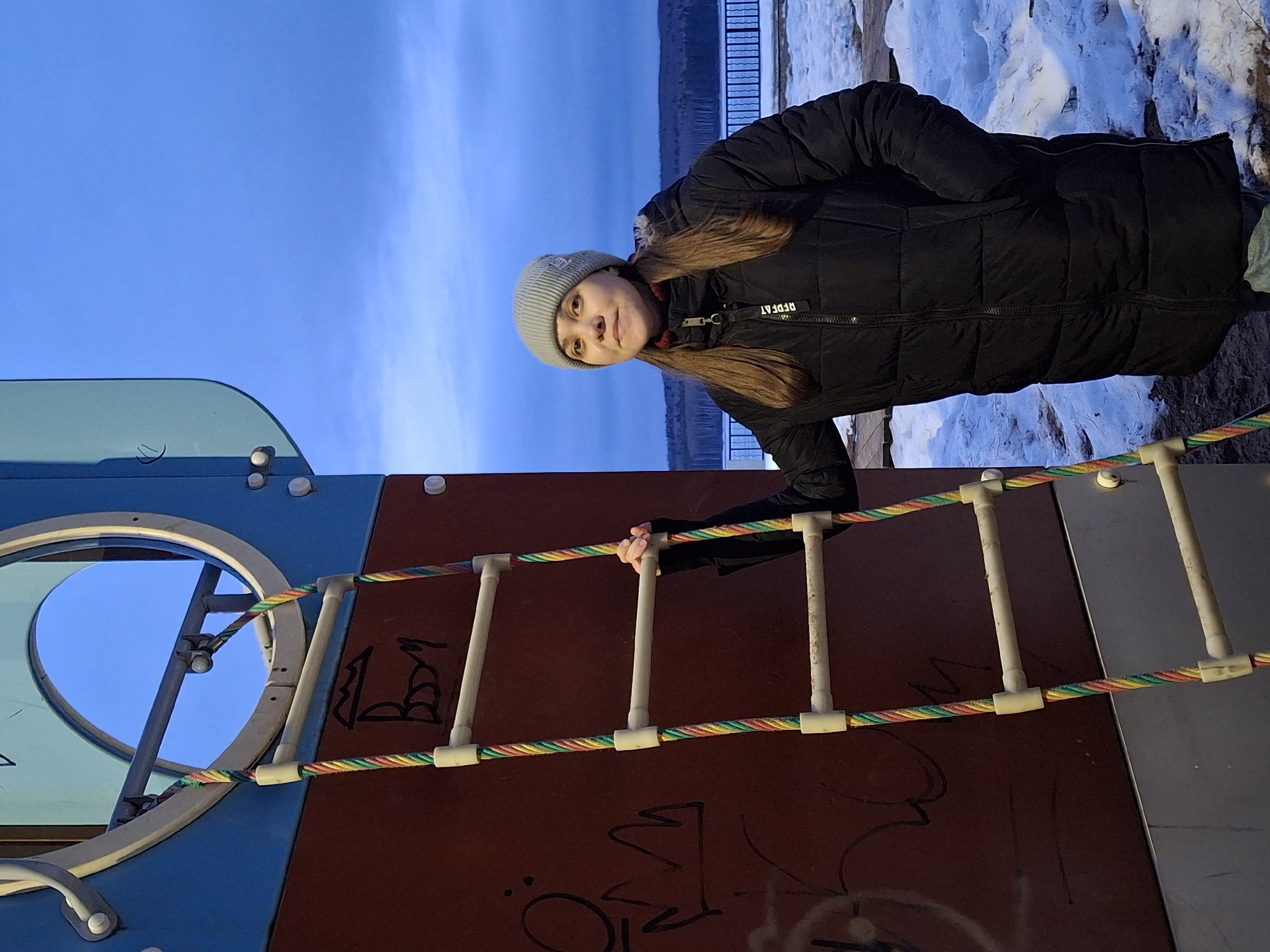 A young woman with long brown hair and a grey beanie is standing on a playground ladder. She is wearing a black puffer jacket. The playground is in Chaikovsky, Russia. The ground is partially covered in snow. The sky is blue. The ladder is attached to a playground structure with blue, brown, and grey panels. There is graffiti on the brown panel.