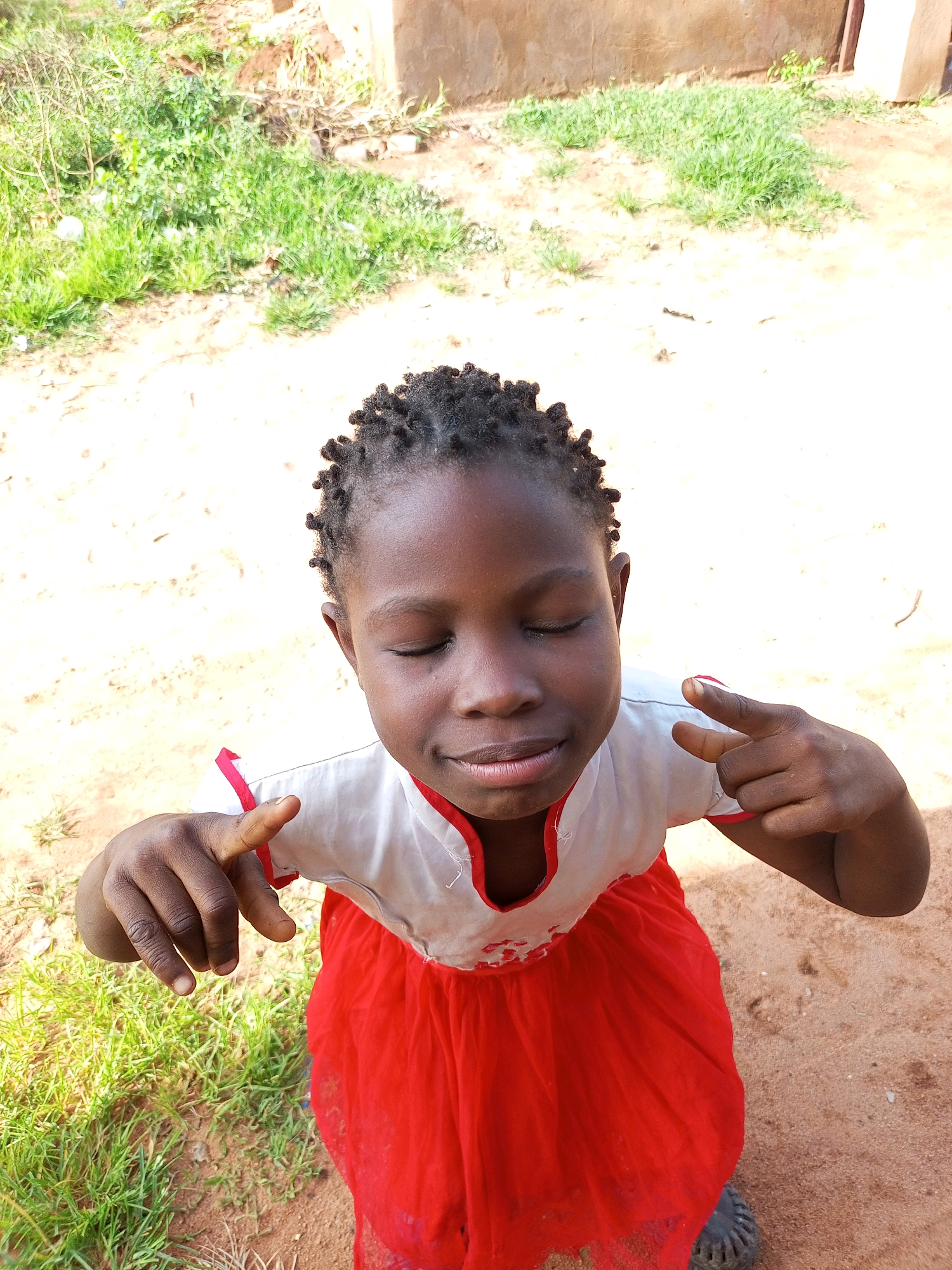 A young girl with braided hair and closed eyes smiles as she holds her hands up in a playful gesture. She is wearing a white top with red trim and a red skirt. The background is a dirt ground with patches of green grass and a building. The image is a high-angle shot, taken outdoors. The lighting is bright, suggesting daytime. The setting appears to be a rural or informal urban environment, consistent with a location in Tempure, Nigeria.