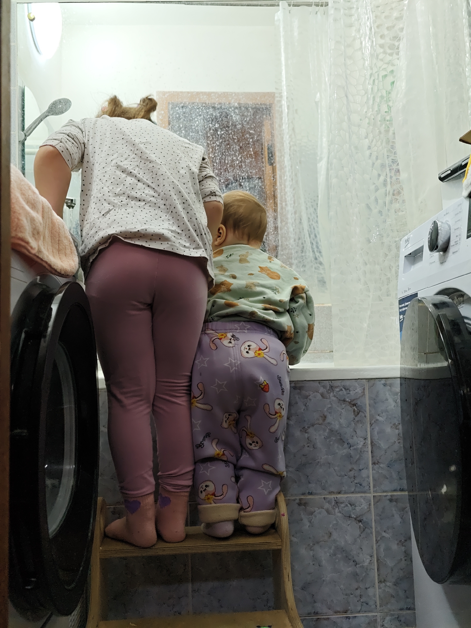 Two children stand on a wooden step stool in a bathroom. The older child wears a white, long-sleeved shirt with black polka dots and pink leggings. The younger child is dressed in a light green, long-sleeved shirt with animal patterns and purple pants with cartoon bunnies. They are both looking into a bathtub, possibly playing with water. A washing machine is visible on either side of them, and a shower curtain hangs along the back wall. The bathroom has blue-grey patterned tiles. The scene is in a bathroom, likely in a residential setting in Chaikovsky, Russia.