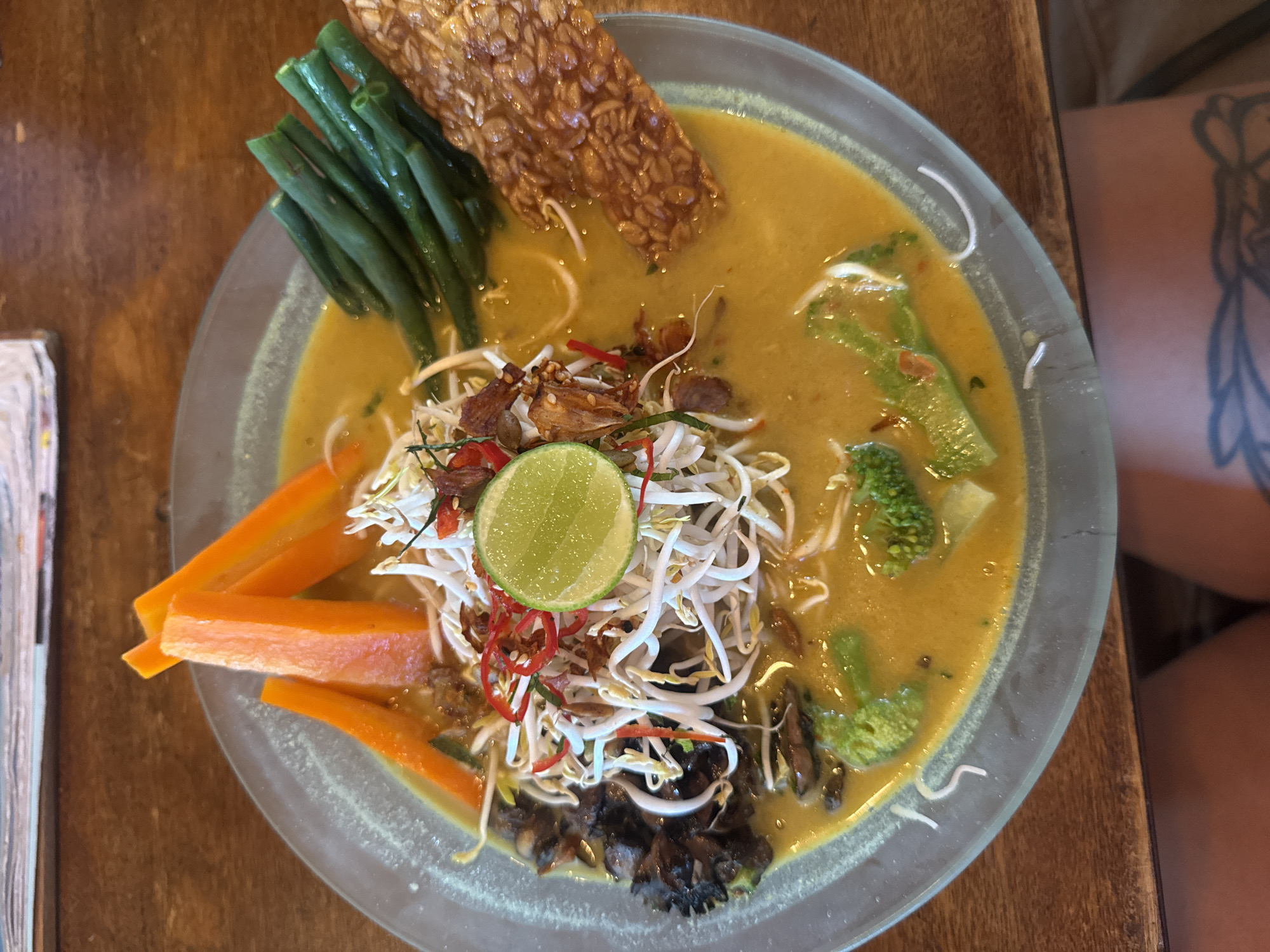 A top-down shot displays a plate of Indonesian vegetarian curry in the Ubud District, Indonesia. The curry is a yellowish-orange color and contains a variety of vegetables. Sliced carrots, green beans, and broccoli florets are visible. A rectangular piece of tempeh sits among the vegetables. A pile of bean sprouts, fried shallots, red chili slices, and a lime slice are in the center. The plate is clear glass with a gray rim and rests on a wooden table. A tattooed arm is visible on the right side of the shot.