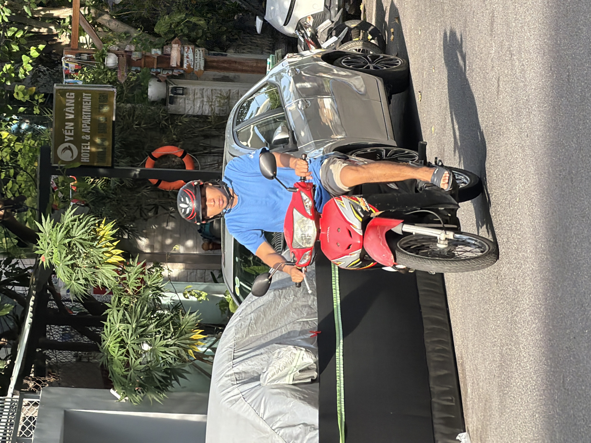 A man wearing a blue t-shirt, khaki shorts, a helmet, and dark sandals rides a red motorcycle. He is on a paved street in Nha Trang, Vietnam, during the day. To his left, a silver car is parked behind a gray vehicle cover. In the background, there is a sign for "YEN VANG HOTEL & APARTMENT" and lush green trees. The man appears to be looking forward, focusing on the road ahead.