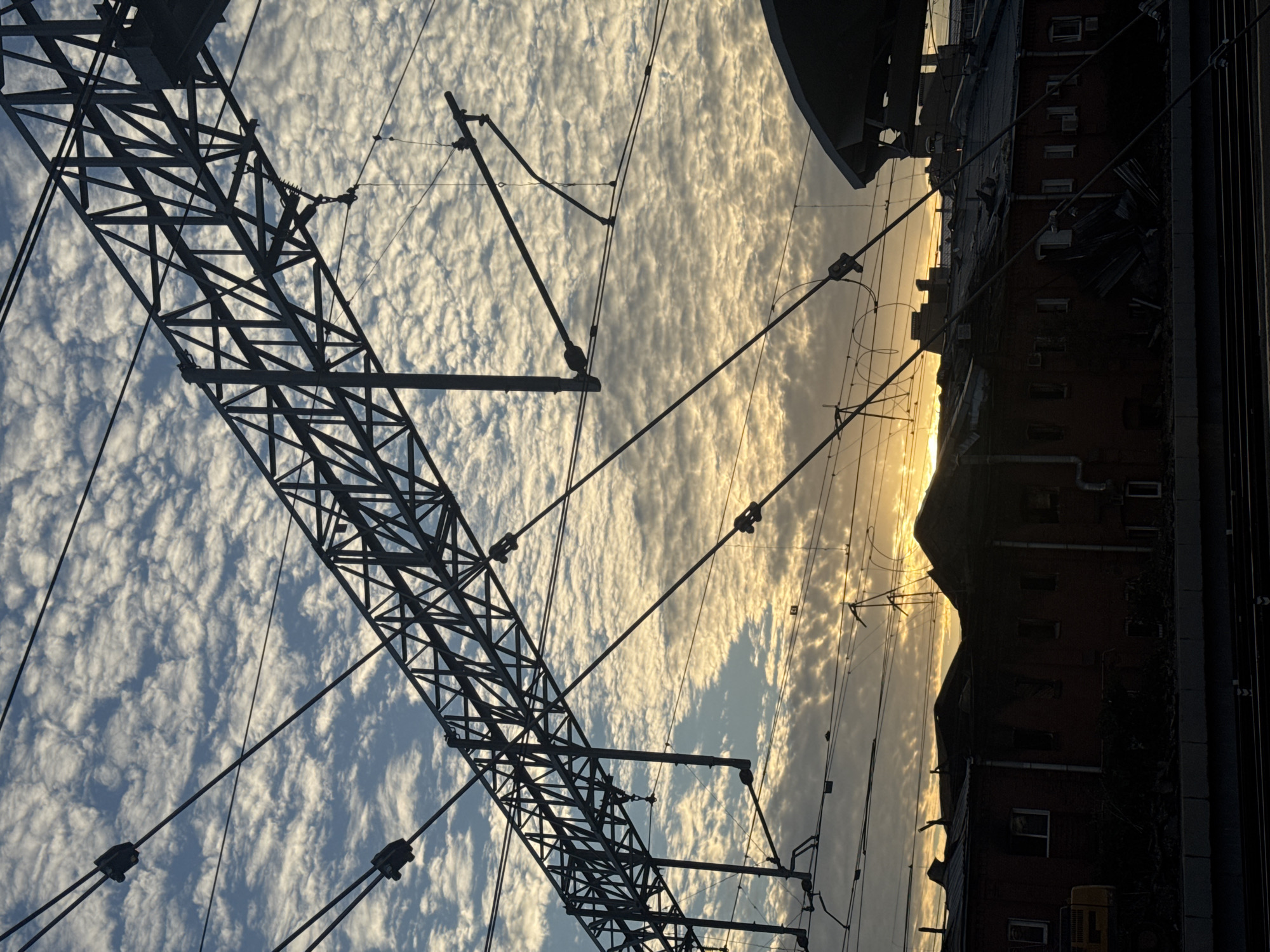 The image depicts a view of a railway line with overhead catenary wires silhouetted against a sky filled with altocumulus clouds. The sun is setting or rising, casting a warm glow on the clouds and silhouetting the industrial infrastructure. In the foreground, a dark, brick building with a damaged roof is visible, suggesting it might be an abandoned or destroyed structure. There is also a yellow excavator partially visible on the left side of the building. The overall scene conveys a sense of urban decay or post-disaster environment. The presence of a railway line and overhead wires suggests this location is within a functional or formerly functional railway station or yard. Without specific landmarks, pinpointing the exact location within Saint Petersburg is not possible from the image alone.