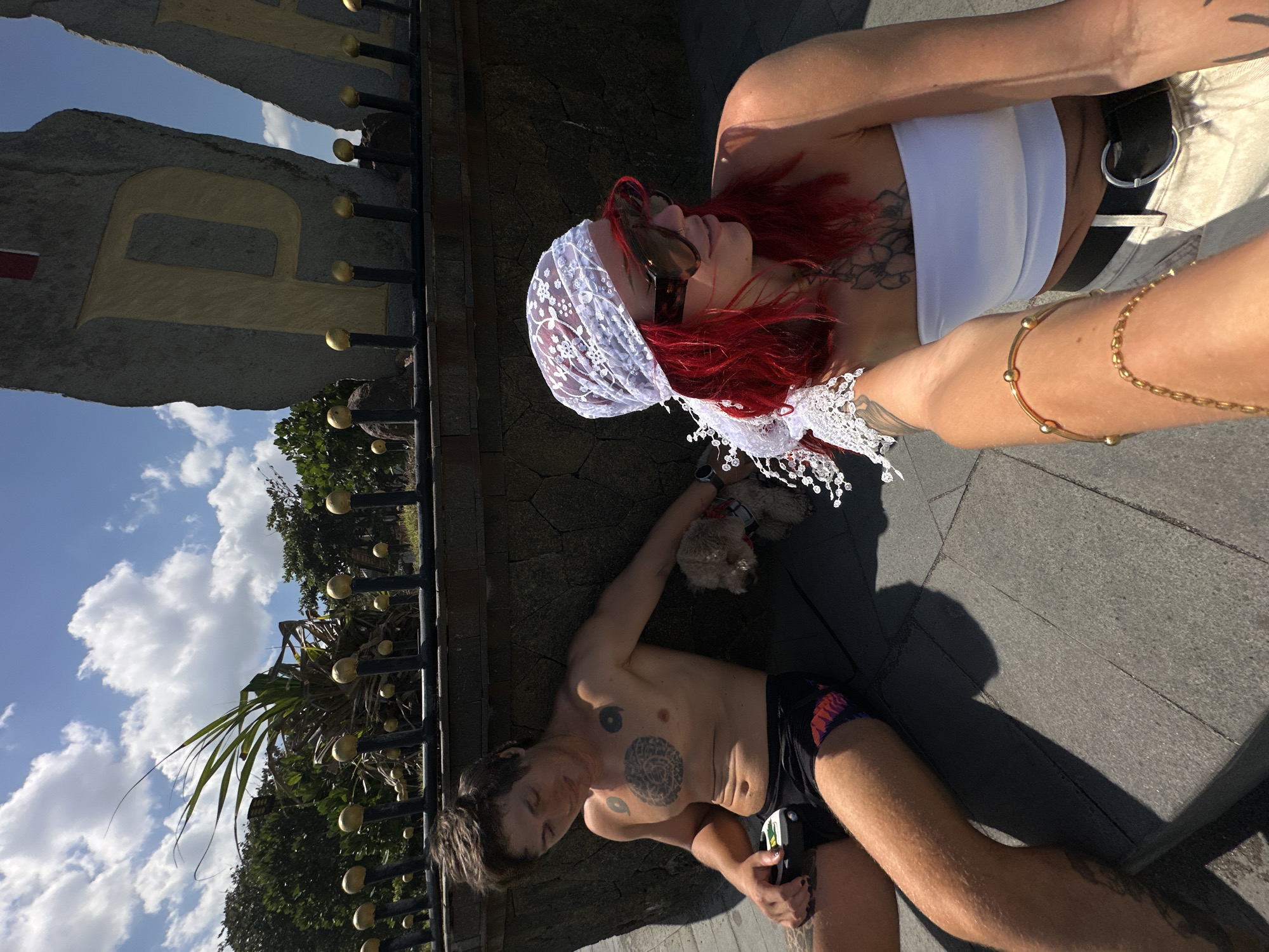 Two individuals and a small dog are resting on a grey paved surface next to a stone wall under a clear blue sky with scattered white clouds in Mengwi, Indonesia.
On the left, a male subject with dark hair, topless and wearing black swim trunks, leans against a textured stone wall. He has a large circular tattoo on his chest and two smaller circular tattoos on his pectorals. He holds a black and green electronic device in his left hand and wears a watch on his left wrist.
Between the two people, a small, light brown fluffy dog wearing a red harness lies on the pavement.
On the right, a female subject with bright red hair, wearing a white lace headscarf and large dark sunglasses, takes a selfie-style photograph. She wears a white strapless top, light-colored pants, and a black belt with a circular buckle. Tattoos are visible on her chest and arm. Her right arm, adorned with multiple gold bracelets, is extended towards the viewer.
Behind them, a dark metal railing with numerous golden spherical finials runs atop the stone wall. Beyond the wall, green trees and a large plant with long, spiky leaves are visible. Further back, two large, rough-hewn stone blocks with the golden letters "P" and "A" carved into them are prominent. A small red and white flag or banner is attached to the top of the "P" block.