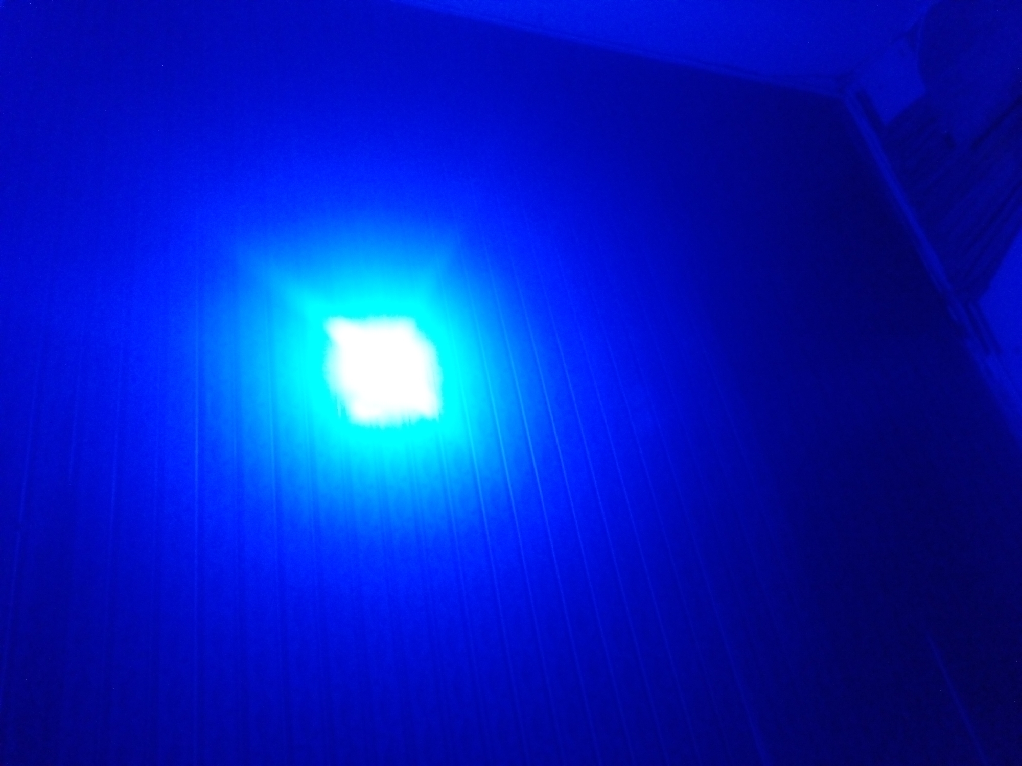 A bright, white light emanates from a square-shaped light source in the center of the image. The light is surrounded by a strong blue glow that illuminates a textured surface with faint, parallel lines. The overall color palette is dominated by deep blues, with the white light creating a stark contrast. The image appears to be taken from a low angle, looking up at the illuminated surface. The environment is dark except for the bright light source.