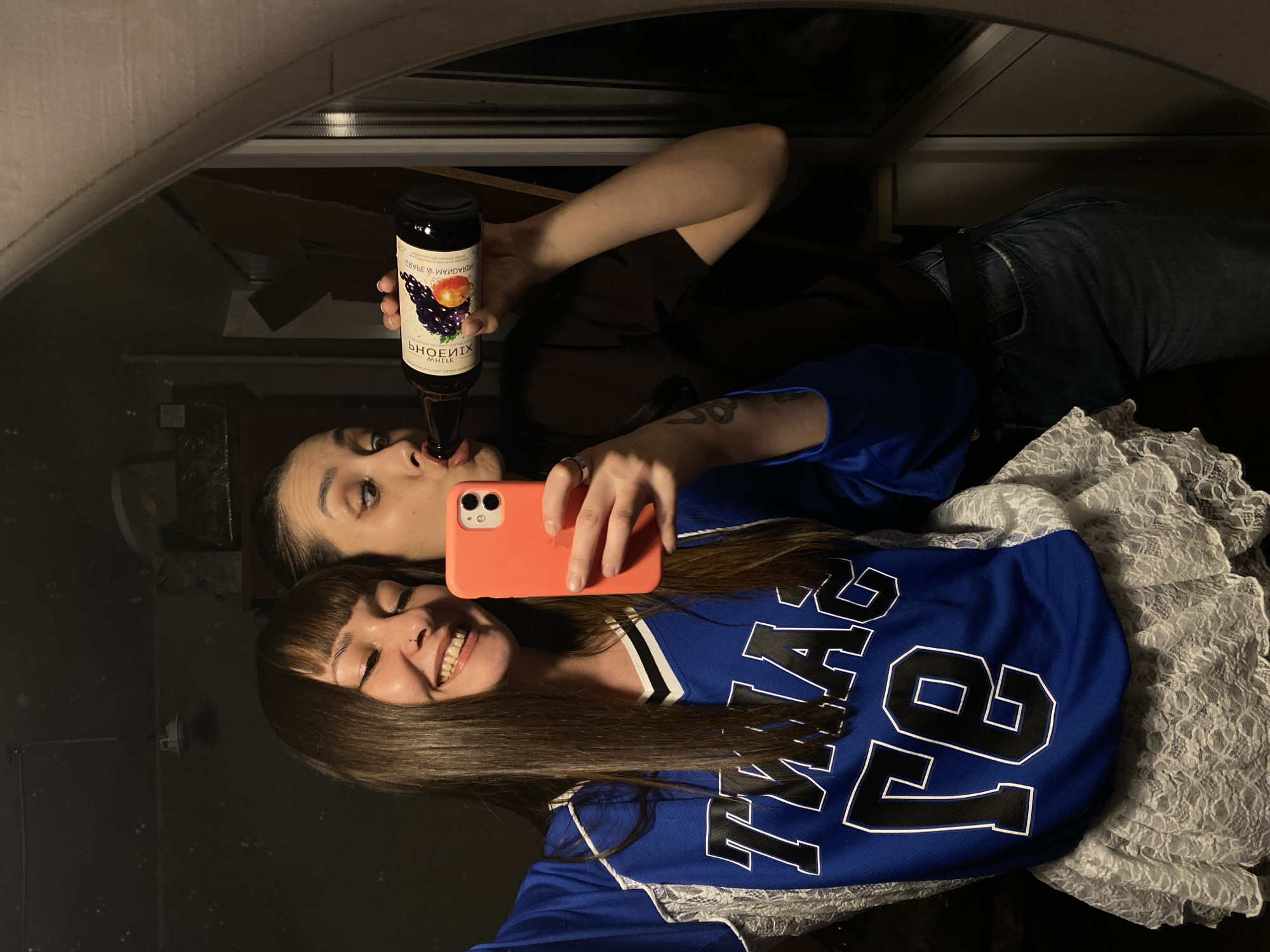 Two young women are taking a selfie in a mirror. The woman in the foreground is smiling, with bangs and long dark hair, wearing a blue "TEXAS 78" jersey over a white lace top. The woman behind her is tilting her head back to drink from a dark glass bottle labeled "PHOENIX MHTLE." She is holding an orange iPhone. The background reflection includes dark furniture. The location of the photo is unknown and not necessarily Votkinsk, Russia.