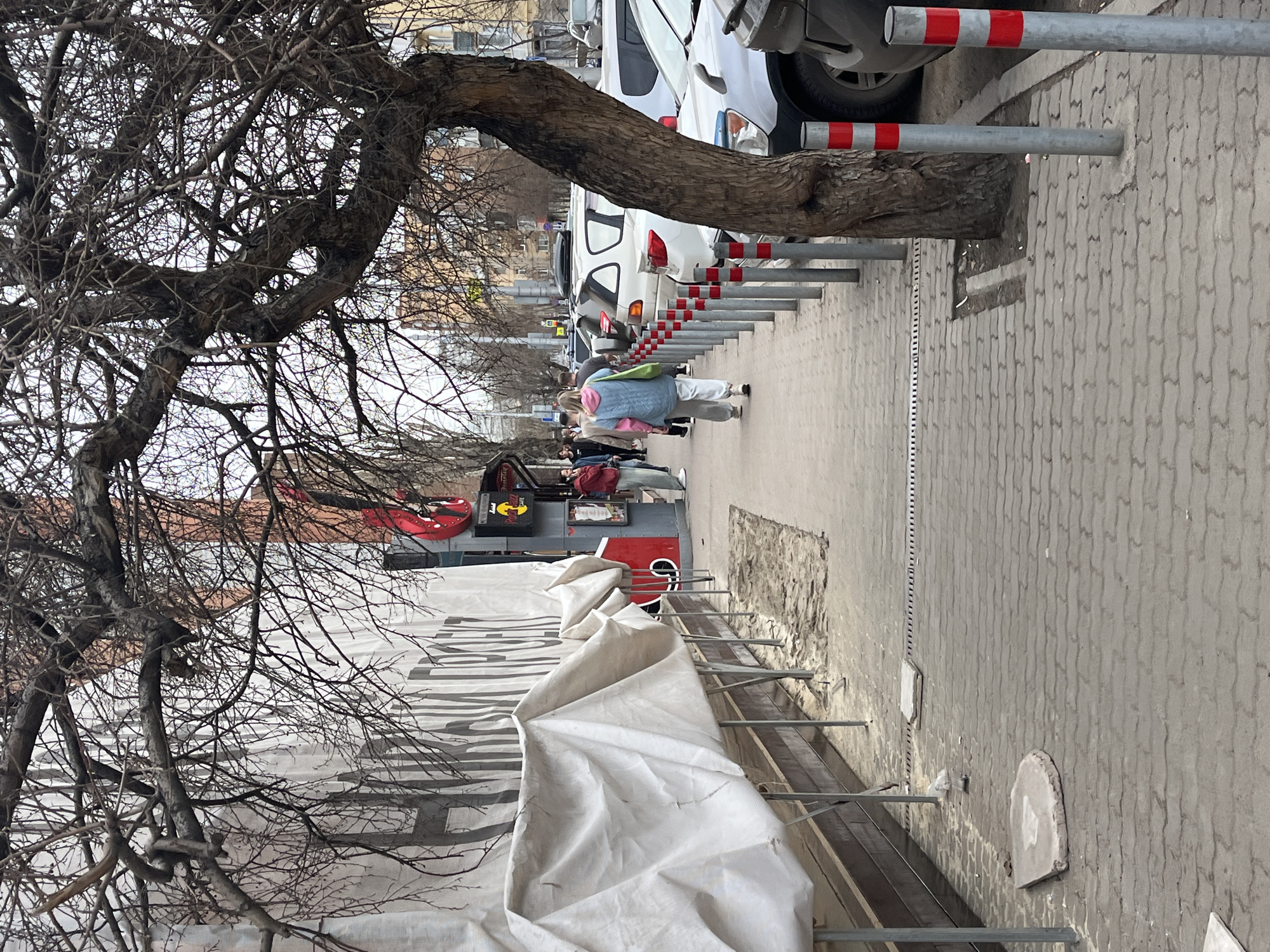 The image shows a street scene in an urban environment. On the right, a sidewalk paved with interlocking blocks runs parallel to a row of closely spaced metal bollards, some with red and white stripes. Beyond the bollards are parked cars. Pedestrians, including a person in a light-blue vest and white pants, are walking along the sidewalk.
To the left, scaffolding covered with white tarps obscures the lower portion of a building. A tree with bare branches is prominently featured in the foreground. Higher up, above the scaffolding, are signs associated with the "Red Bull" brand, partially obstructed by the tree. The sky appears overcast.