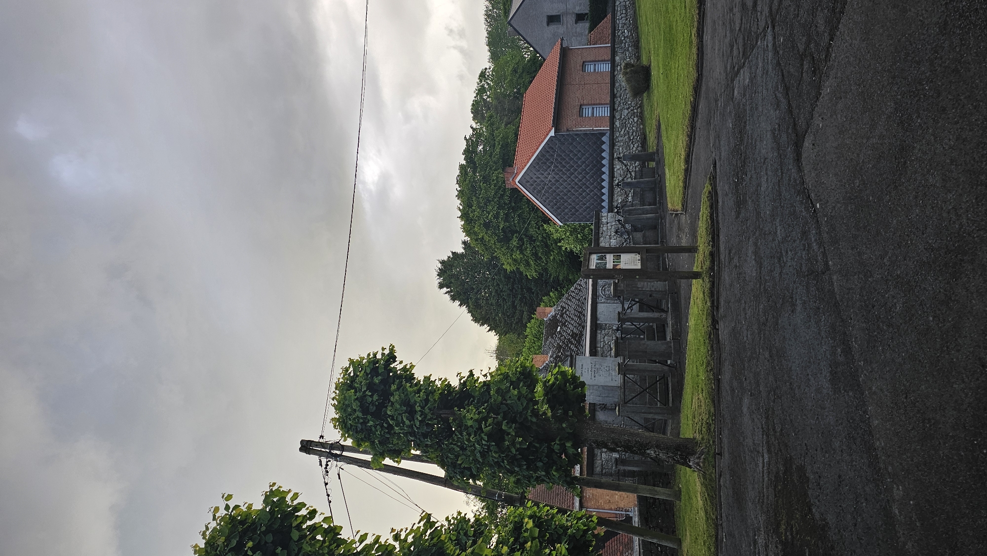 This image captures a tranquil village scene in Boussu-lez-Walcourt, Belgium, under an overcast sky, suggesting late morning or early afternoon after a recent rain. The ground is dominated by a wet, dark asphalt road, showing cracks, that recedes into the background on the right side of the frame.
To the left of the road, a narrow strip of vibrant green grass lines the path, backed by low, grey stone walls in places. In the mid-ground, a section appears to be a small churchyard or cemetery, featuring several weathered grey grave markers or monuments. A rustic wooden fence or gate structure and a small signpost are also present in this area; any text on the sign or monuments is unreadable due to distance and blurriness.
A tall, slender tree with dense green foliage stands prominently in the mid-ground on the left, next to a utility pole with wires extending across the sky. In the background, traditional Belgian houses with distinct architectural features are visible. One notable house has red brick walls, a steeply pitched red tiled roof, and a unique dark, patterned section on its gable end. Further back and to the left, another house with a grey roof and grey walls is partially visible, nestled among dense green trees that define the horizon. The sky is entirely covered with grey, heavy clouds, with a slightly brighter area indicating the sun's obscured position. The scene is devoid of people or discernible activity, conveying a quiet, serene, and somewhat melancholic atmosphere.