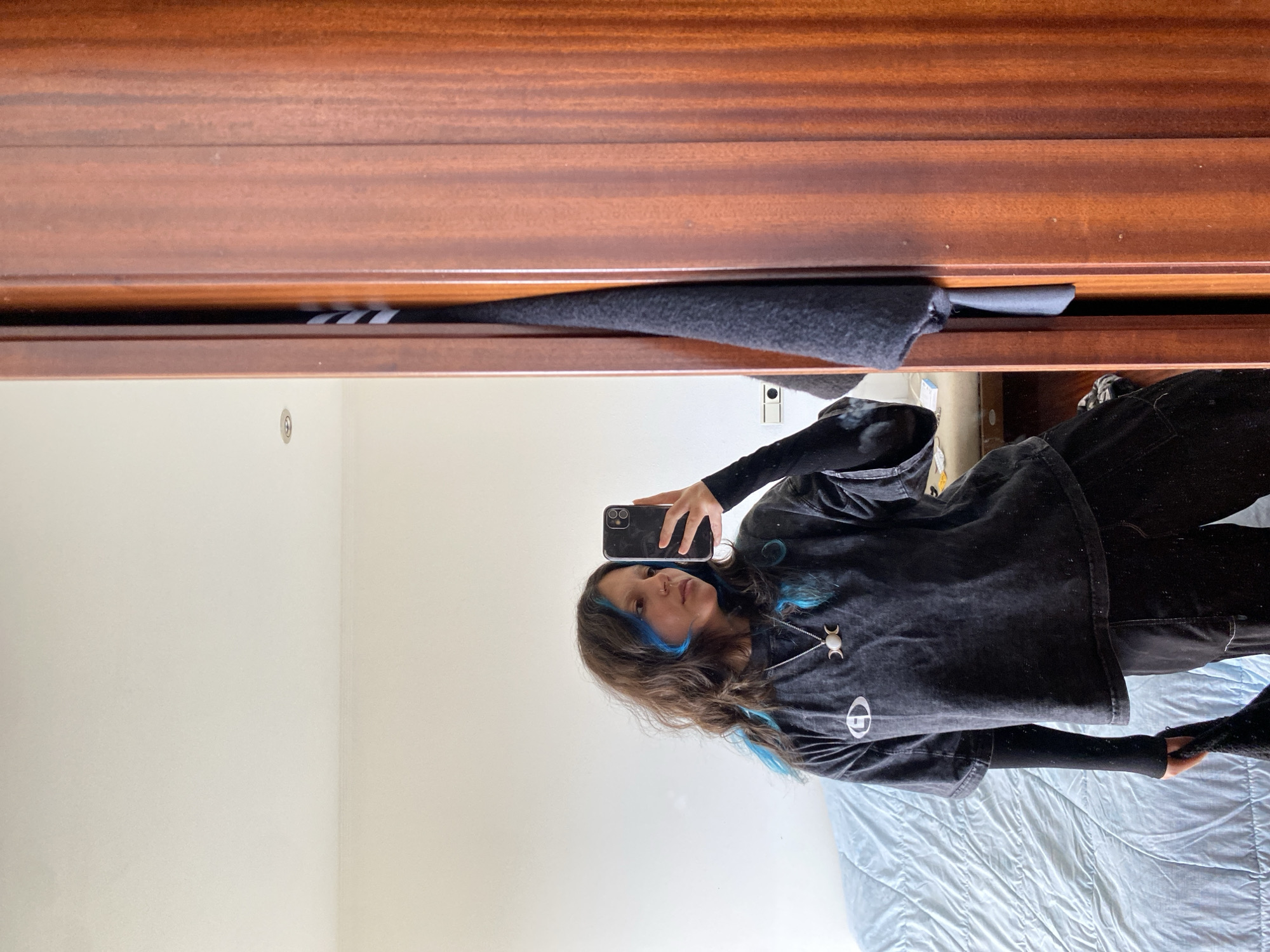 A woman is reflected in a mirror, holding a cell phone up to take a photo of herself. She has long brown hair with streaks of bright blue, a dark shirt with a silver necklace, and black sleeves. A portion of a wooden dresser, presumably the mirror’s frame, is visible along the top edge of the image. The background features a plain white wall and a light blue bedspread. The scene does not reveal information about a specific location.