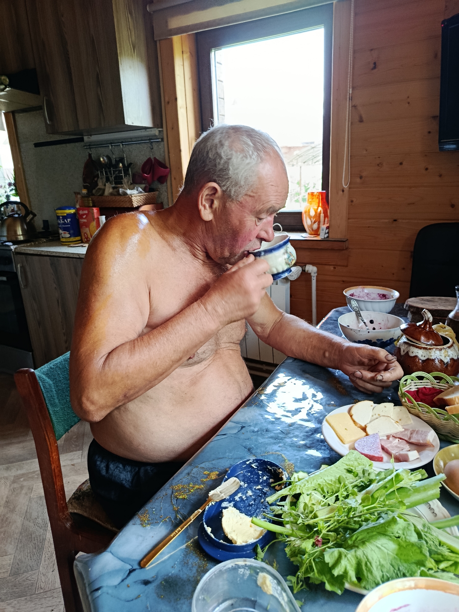 An elderly, shirtless man with graying hair is seated at a table, drinking from a patterned cup. He is indoors, in what appears to be a rustic kitchen or dining area with wood-paneled walls. On the blue-covered table are various breakfast items, including bowls of berries and yogurt, slices of cheese and ham on a plate, a basket of bread, a dish of butter, and a large pile of leafy greens. The man's skin appears to be tanned and slightly oily. A window with natural light streaming in is visible behind him. The scene is consistent with a meal being prepared or consumed in a rural setting, possibly a dacha, in Verea, Russia.