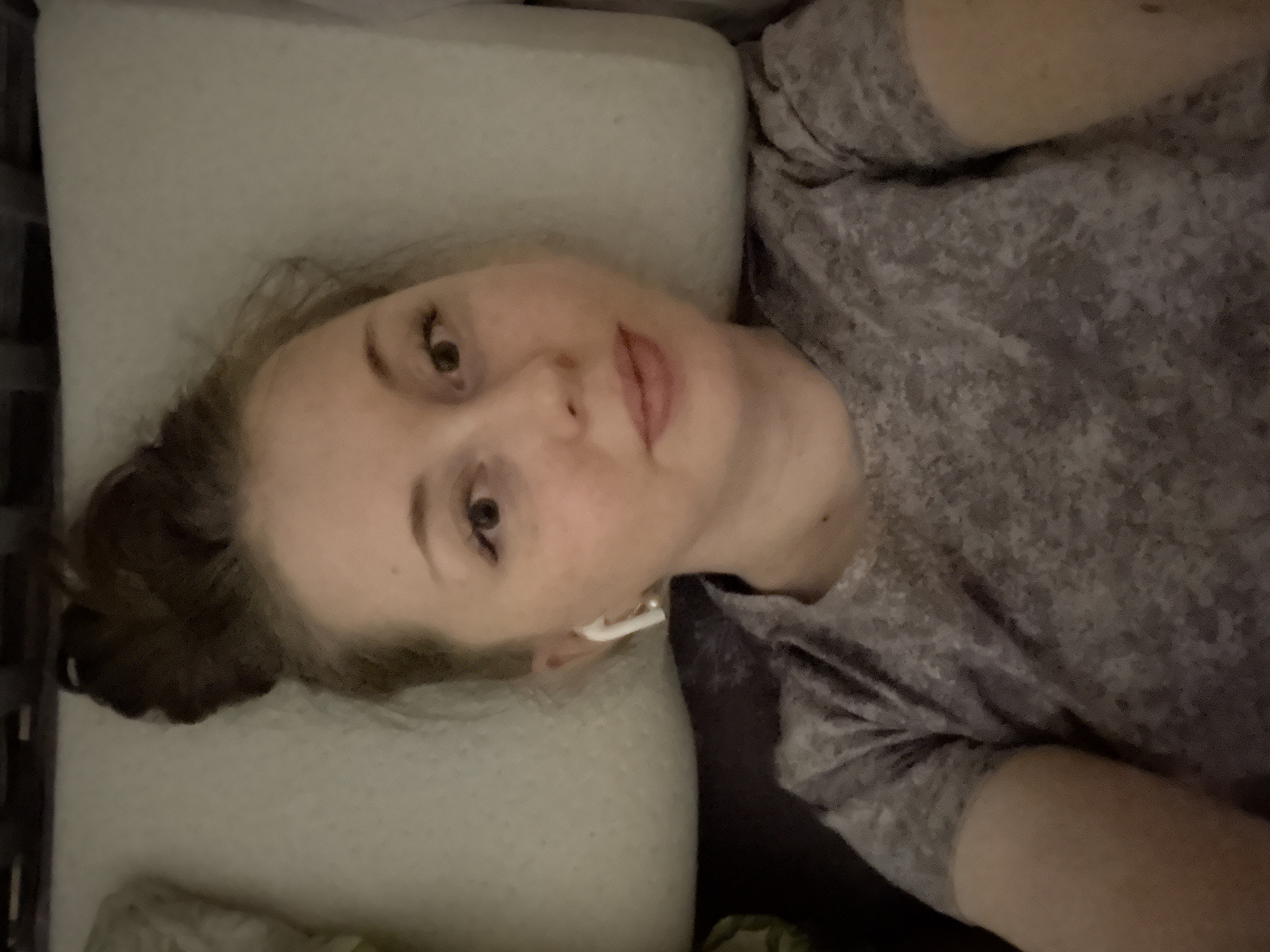 A woman is lying on a light-colored pillow in what appears to be a bed. She has light skin, brown hair pulled into a bun, and is wearing a gray t-shirt. An earbud is visible in her right ear. The location is Unknown, Unknown.