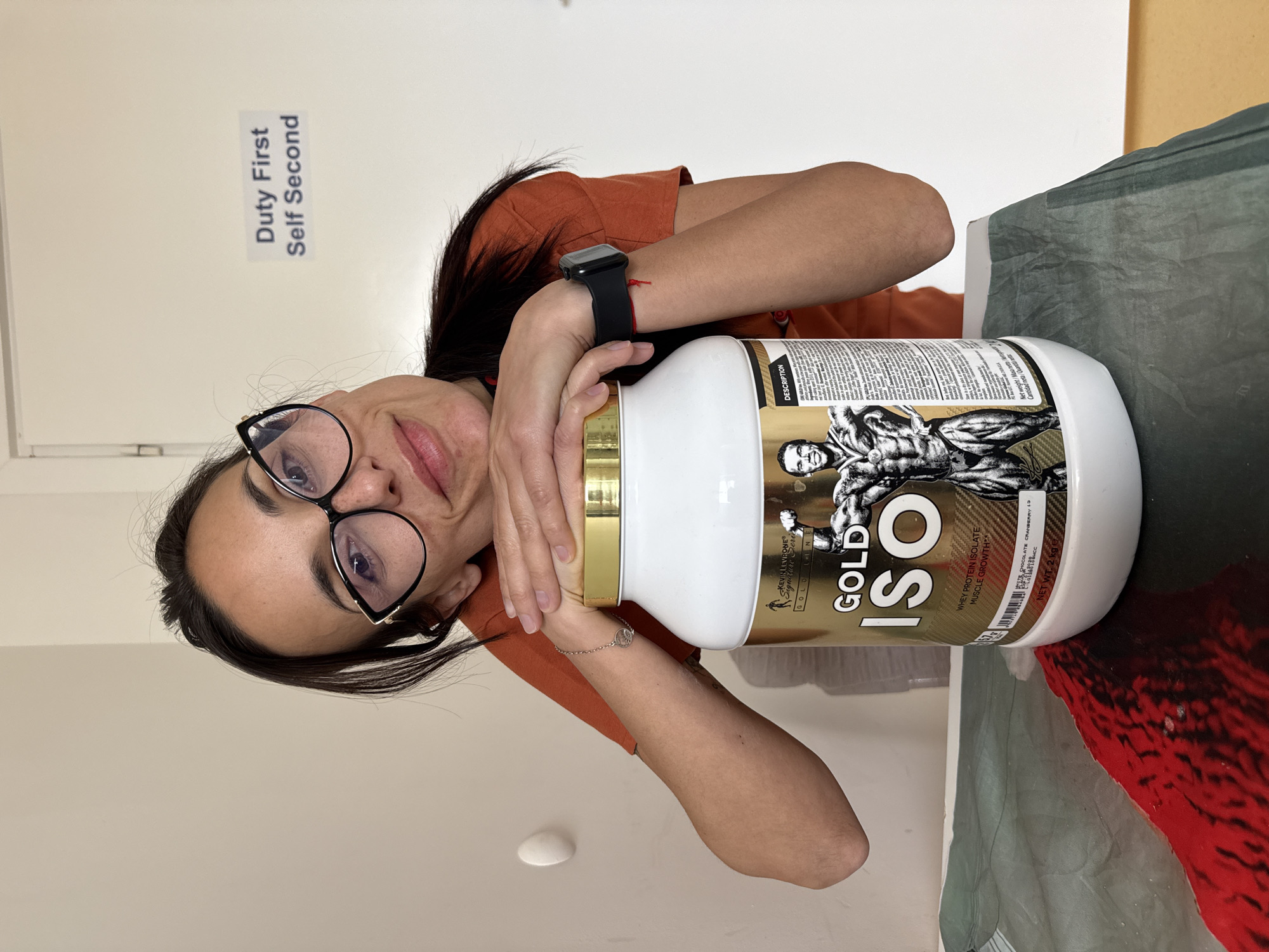 A woman with dark hair and glasses holds a large container of Kevin Levrone Gold Iso whey protein isolate against her chest. She wears an orange shirt and a black smartwatch. A sign above her reads "Duty First Self Second." The container is white with a gold lid and features an image of a muscular bodybuilder. The whey protein is white chocolate cranberry flavor and the net weight is 2 kg. The background consists of a white wall and a surface covered in green and red fabric. The location is implied to be Pleven, Bulgaria.