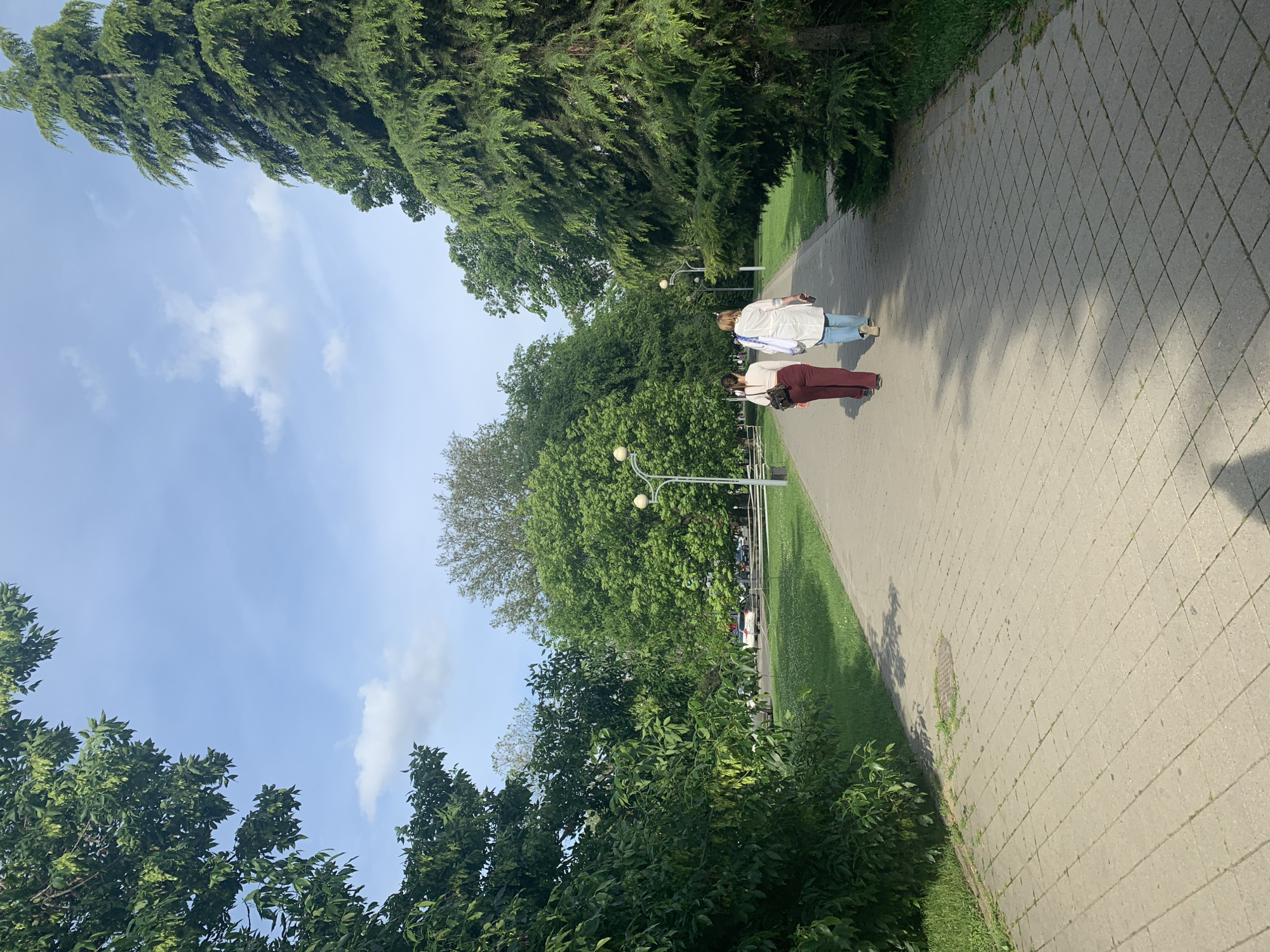 The image captures a paved pathway lined with lush greenery and trees, under a partially cloudy sky. Two people are walking away from the viewer on the path: one wearing a white top and jeans, and the other a white top and maroon pants. Street lamps are visible along the path. The scene appears to be a park or garden. The location is Unknown, Unknown.