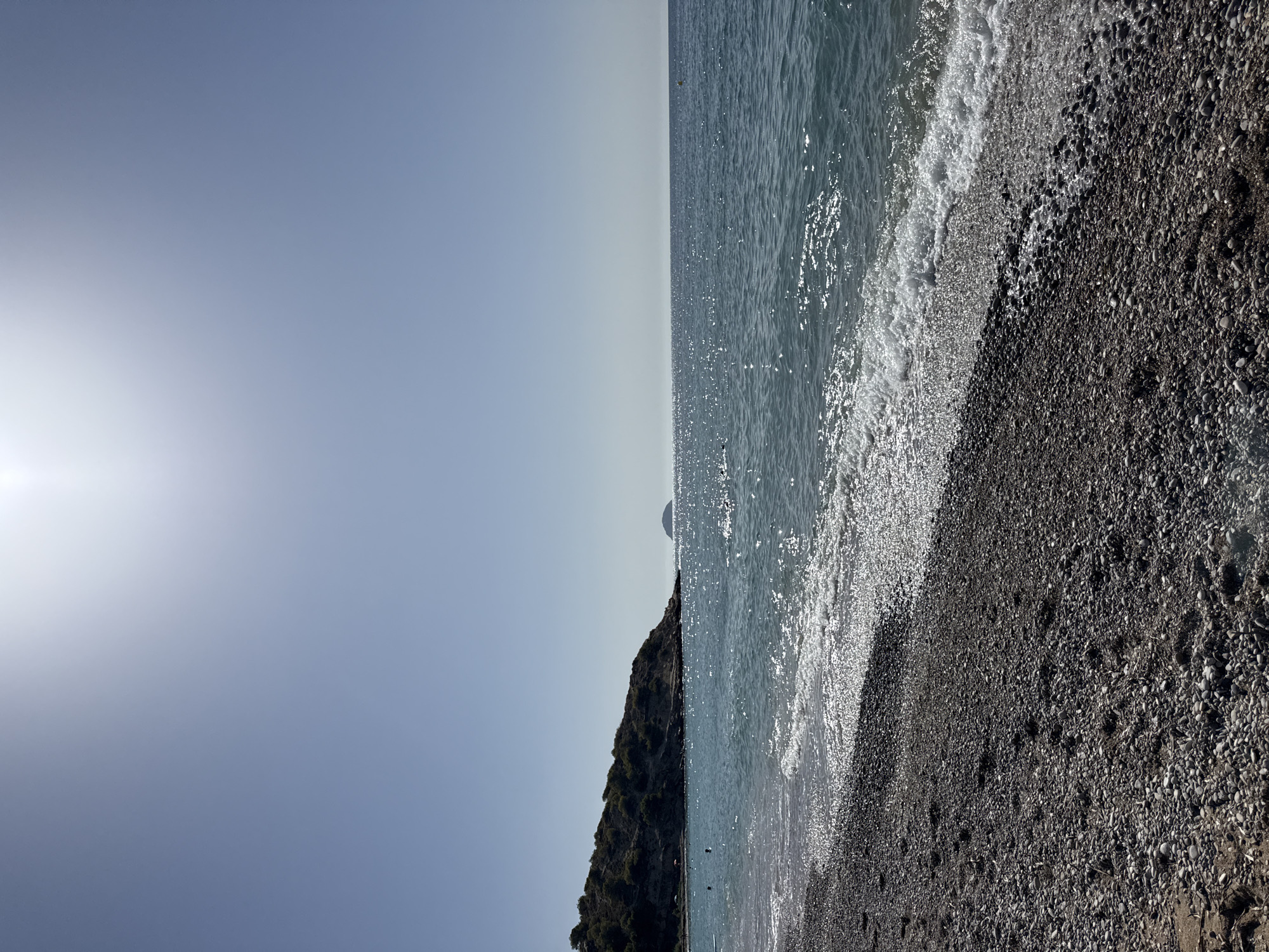 The image depicts a bright, sunny day at a pebble beach in La Vila Joiosa, Spain. The foreground is dominated by a dark pebble beach with some scattered sand. Gentle waves with white foam lap at the shore. In the middle ground, the clear turquoise water shimmers with sunlight. A tree-covered hill with a rugged coastline forms the backdrop on the left side of the image, with a few distant figures visible near the water. In the distance, a small, isolated island can be seen in the middle of the sea. The sky is a clear, pale blue, with the sun high and bright, casting a glare. A yellow buoy is visible in the water towards the right side of the frame.