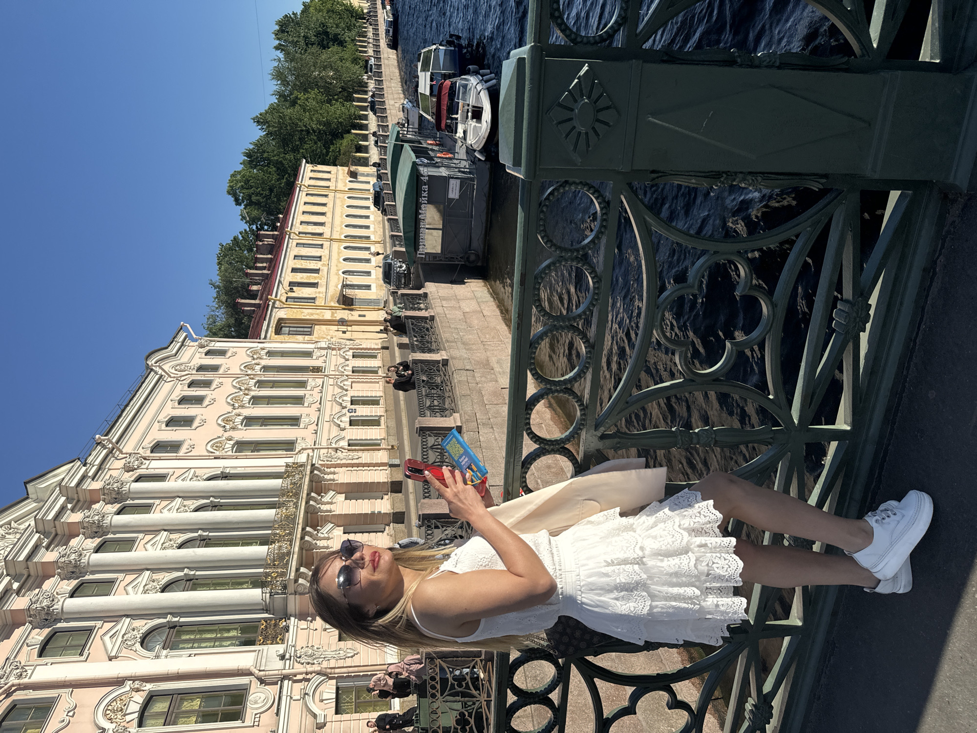 A woman stands on a bridge in Saint Petersburg, Russia, facing to the right and looking at her phone. She is wearing a white, tiered dress and white platform sneakers. She holds a smartphone in her right hand and a small object, possibly a card or ticket, in her left hand. Behind her, a canal runs alongside buildings with classical architecture. Several boats are docked on the canal, and trees line the opposite bank. The sky is clear and blue. The woman is on the Palace Bridge (Dvortsovy Most), which spans the Neva River, connecting to the Admiralty Island and Vasilievsky Island. The building visible in the background on the left is likely the Hermitage Museum or the General Staff Building.