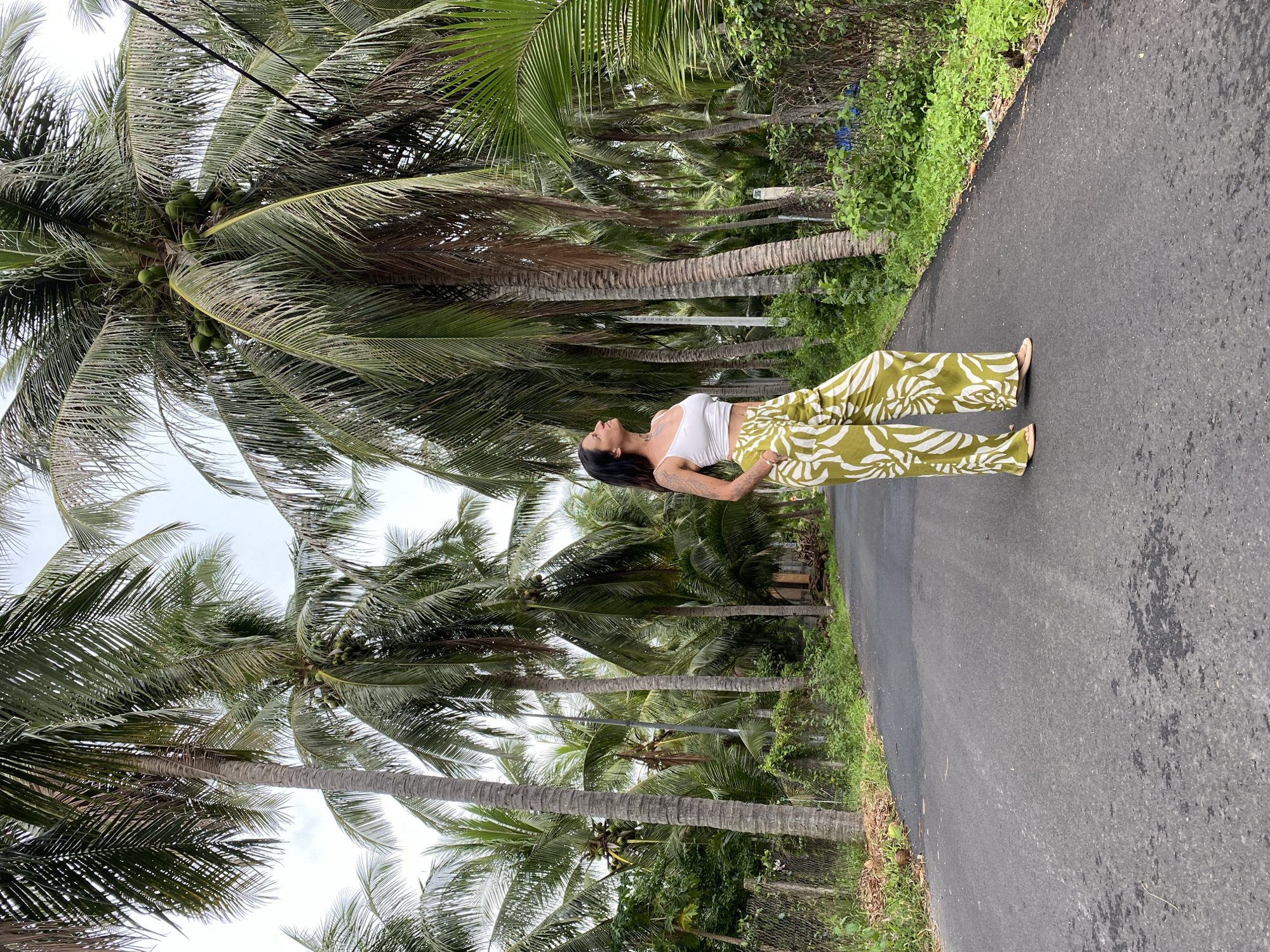 In Mũi Né, Vietnam, a woman with dark hair and tattoos on her arm stands on an asphalt road surrounded by numerous tall coconut trees. She is wearing a white crop top and loose-fitting green and white patterned pants. Her head is tilted upwards, and she is looking towards the sky. The road curves slightly to the right and is lined with lush green vegetation and a wire fence on the left side. The sky is overcast.