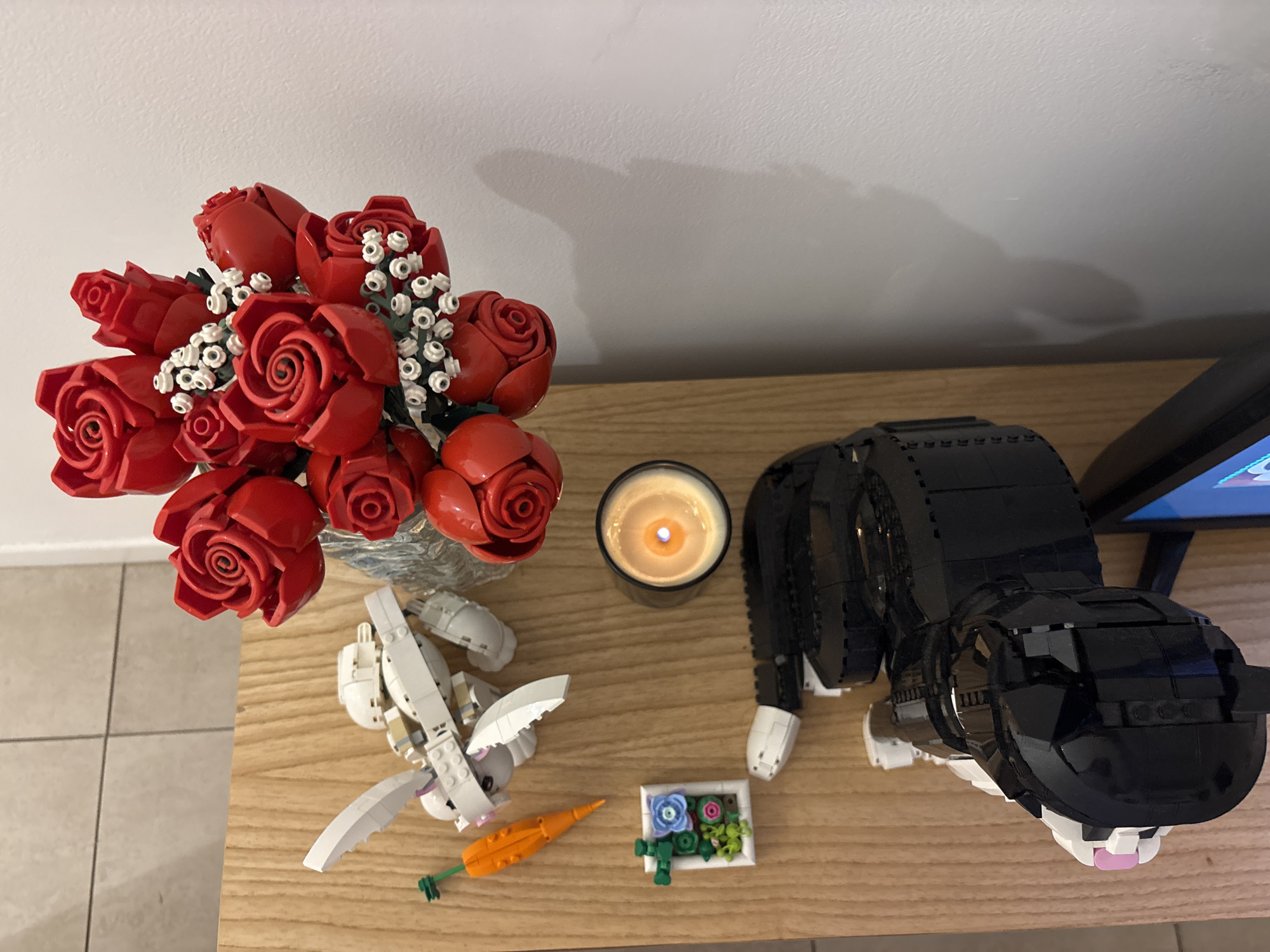 A high-angle shot depicts a collection of objects arranged on a light-brown wooden surface, likely a table or countertop. The scene includes a bouquet of red roses and white flowers in a transparent vase. Adjacent to the flowers is a lit candle in a dark container. A white LEGO bunny with an orange LEGO carrot and a LEGO frame with blue and pink flowers are placed nearby. A LEGO black cat figure is positioned to the right of the candle. The location is not evident from the image, and there is no indication that it is in Buenos Aires, Argentina.