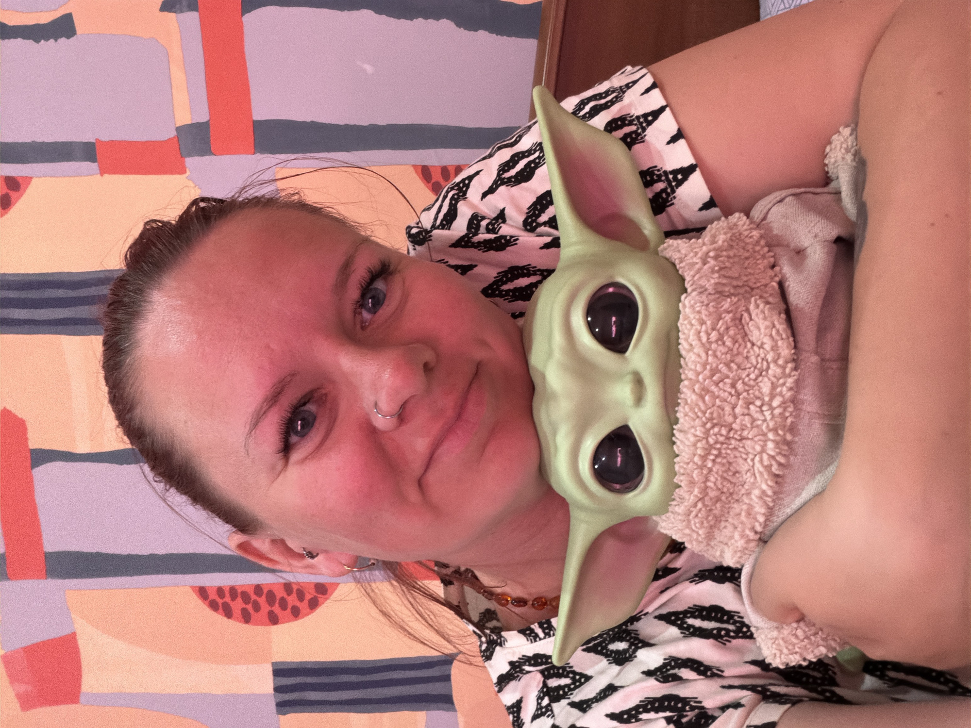 A woman is holding a plush Baby Yoda toy. The woman has her hair pulled back, blue eyes, and is wearing a black and white patterned shirt. She has a nose ring and is wearing a necklace with amber beads. The Baby Yoda plush toy is wrapped in a tan, fuzzy blanket. The background is a wall with an abstract design of various colors, including peach, lavender, grey, and coral. The location of the image is unknown and unrelated to Chaikovsky, Russia.